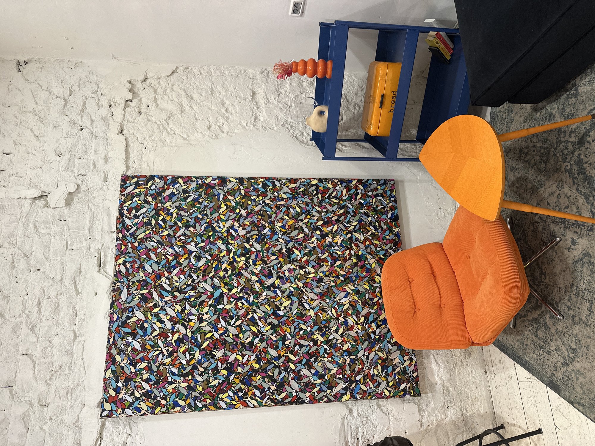 The image shows an interior scene with an exposed brick wall, painted white, acting as the backdrop. A framed, colorful piece of art adorns the wall. Adjacent to the artwork is an orange chair, and a blue shelf filled with a bread holder. The scene seems to be indoors, based on the architectural features. This picture was most likely taken in Tel Aviv, Israel due to architectural styles and the context clues provided by the objects inside.