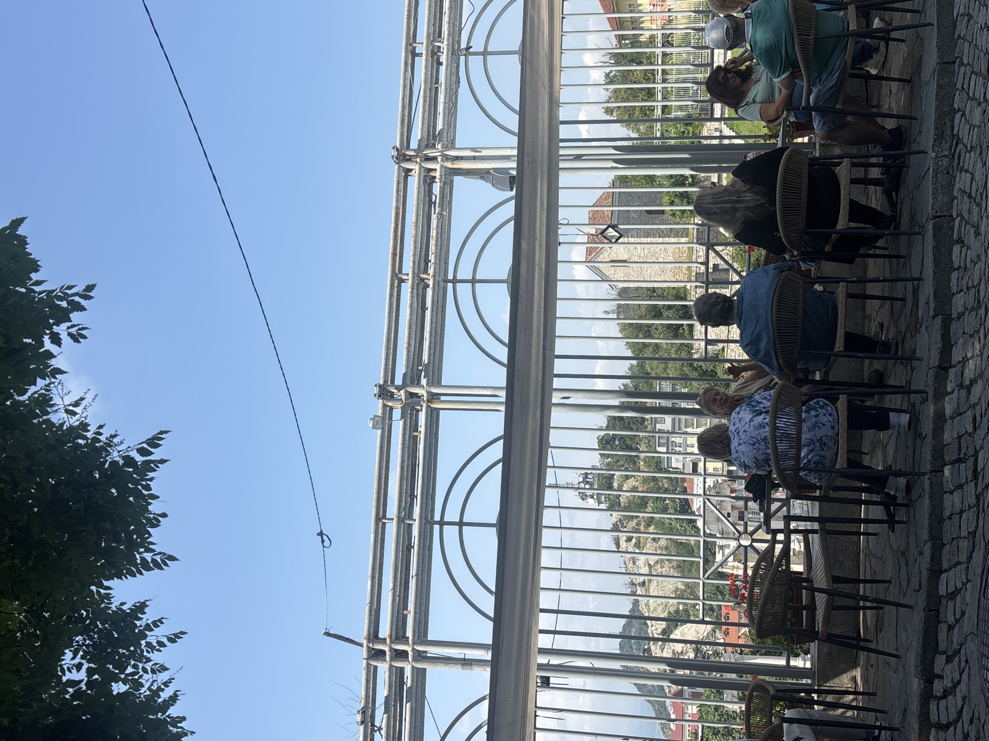 An outdoor scene depicts several individuals seated at tables, likely at a café or restaurant with a scenic view, in Plovdiv, Bulgaria. The image is taken from a slightly elevated position, looking across a metal balustrade or fence with vertical bars and semicircular decorative arches.
On the right side of the image, which appears to be the foreground, the ground is covered in cobblestones. Several people are seated on chairs with wicker-style backs and dark frames, around small tables. Most of the individuals are seen from behind or partially, engaged in what appears to be casual conversation or dining. One person wears a light blue patterned shirt, another a dark top with long grey hair, and further along, individuals are seen in a dark blue shirt, a light green top, and a white hat or helmet.
Beyond the metal fence, a landscape unfolds with lush green trees and foliage, various buildings with reddish-brown tiled roofs, and distant rocky hills or mountains. The sky is clear and bright blue, suggesting a sunny day, likely during late morning or afternoon. A thin, dark cable stretches across the sky, with a small loop or hook visible on it. The overall atmosphere appears relaxed and leisurely. No visible text is present in the image.