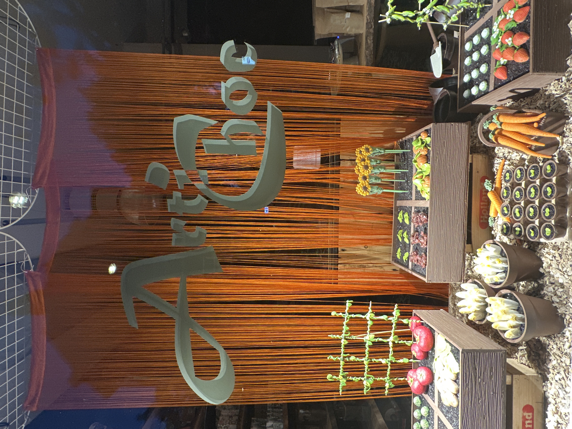The image shows a chocolate display window, likely from a store named "Art'Choc" as indicated by the prominent lettering. The display is designed to resemble a garden, with various chocolate confections representing vegetables, fruits, and plants.
In the foreground, there are chocolate carrots in a basket, and rows of small chocolate sprouts or seedlings in pots. To the left, there are chocolate apples and what appear to be chocolate bananas in a planter box, with a green lattice structure above. In the center and right, there are planter boxes containing chocolate representations of lettuce, pumpkins, and strawberries. Two brown pots hold chocolate asparagus or similar plant-like confections. Behind the planters, an orange string curtain hangs, and a white grid structure is visible at the top of the display.
The setting appears to be a retail environment, likely a chocolatier or confectionery shop. The presence of wooden crates labeled "Holland" suggests a connection to Dutch produce or goods. The overall impression is a creative and artisanal presentation of chocolate products.
While the image itself does not explicitly state the location, the presence of "Holland" on the crates strongly suggests a Dutch context. If this display is indeed from the store "Art'Choc," a search for that establishment within Amsterdam, Netherlands, would be necessary to confirm the specific location. Without further information or external data, it is not possible to pinpoint the exact street or address in Amsterdam.