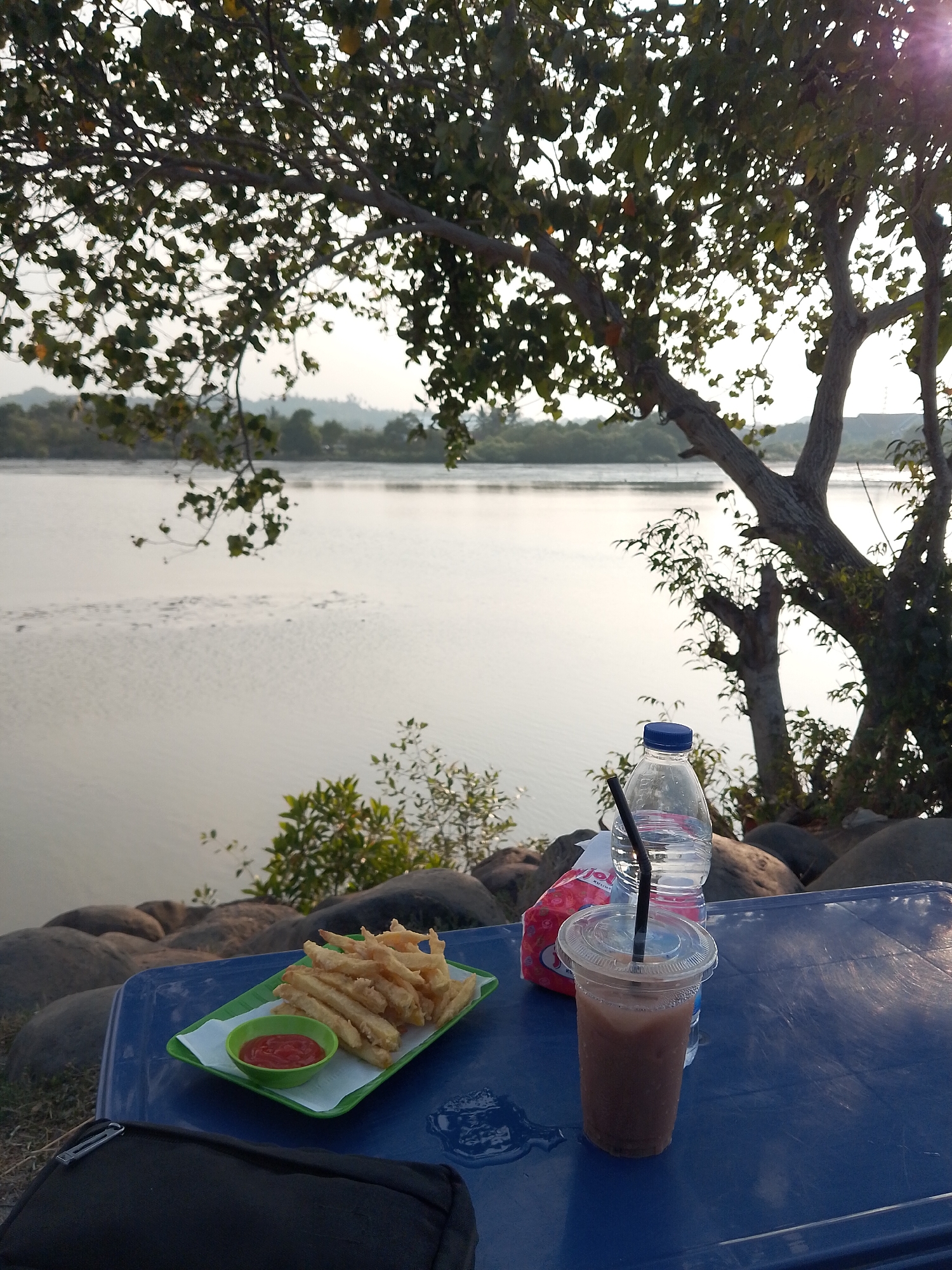 The image depicts an outdoor setting by a body of water, likely a river or sea, with trees and vegetation in the background. In the foreground, a blue table holds several items. These include a green rectangular plate of french fries with a small bowl of ketchup, a clear plastic cup with a brown beverage and a black straw, and a bottle of water with a blue cap. Behind the bottle of water, a folded packet of tissues is visible. The ground around the table is covered with large, dark rocks. The lighting suggests it is either early morning or late afternoon. The location is described as being in Lhokseumawe, Indonesia. There are no people visible in the image.