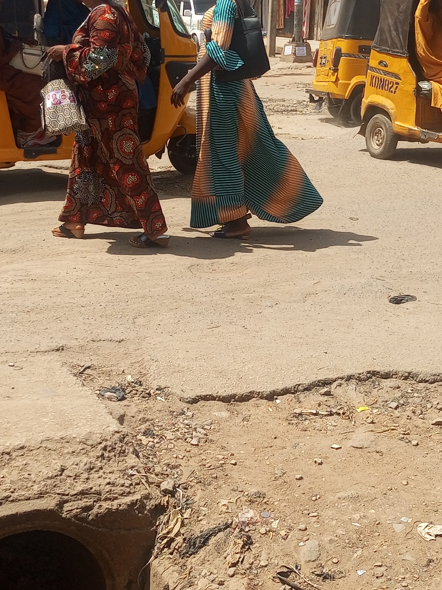 The image captures a street scene in Maiburiji, Nigeria. Two women wearing long, colorful dresses are walking on a paved road that shows signs of wear and tear. One woman's dress is patterned with red, brown, and orange circles, while the other's dress features green and orange stripes. Yellow auto-rickshaws, locally known as keke, are visible in the background. The foreground shows a damaged road surface with exposed soil and a partially visible drain or sewer opening.