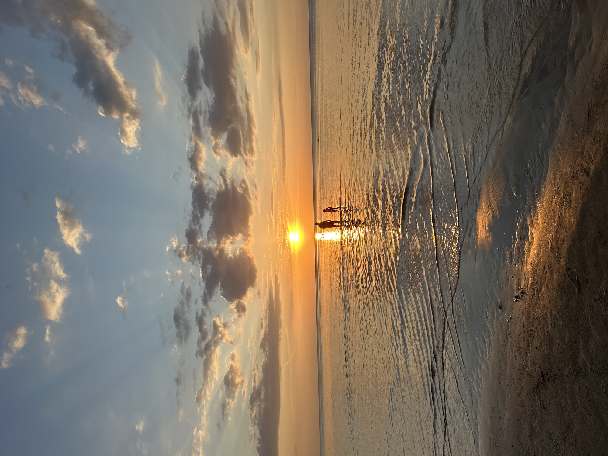 The image shows a sunset over the Baltic Sea, viewed from the beach in Jūrmala, Latvia. The sun is low on the horizon, casting a golden glow across the water and the sky. Several clouds are scattered across the sky, some illuminated by the sun's rays. Two people, a larger silhouette and a smaller silhouette, are standing in the shallow water near the shore. The water is calm, with small ripples reflecting the light. The sandy beach is visible in the foreground.