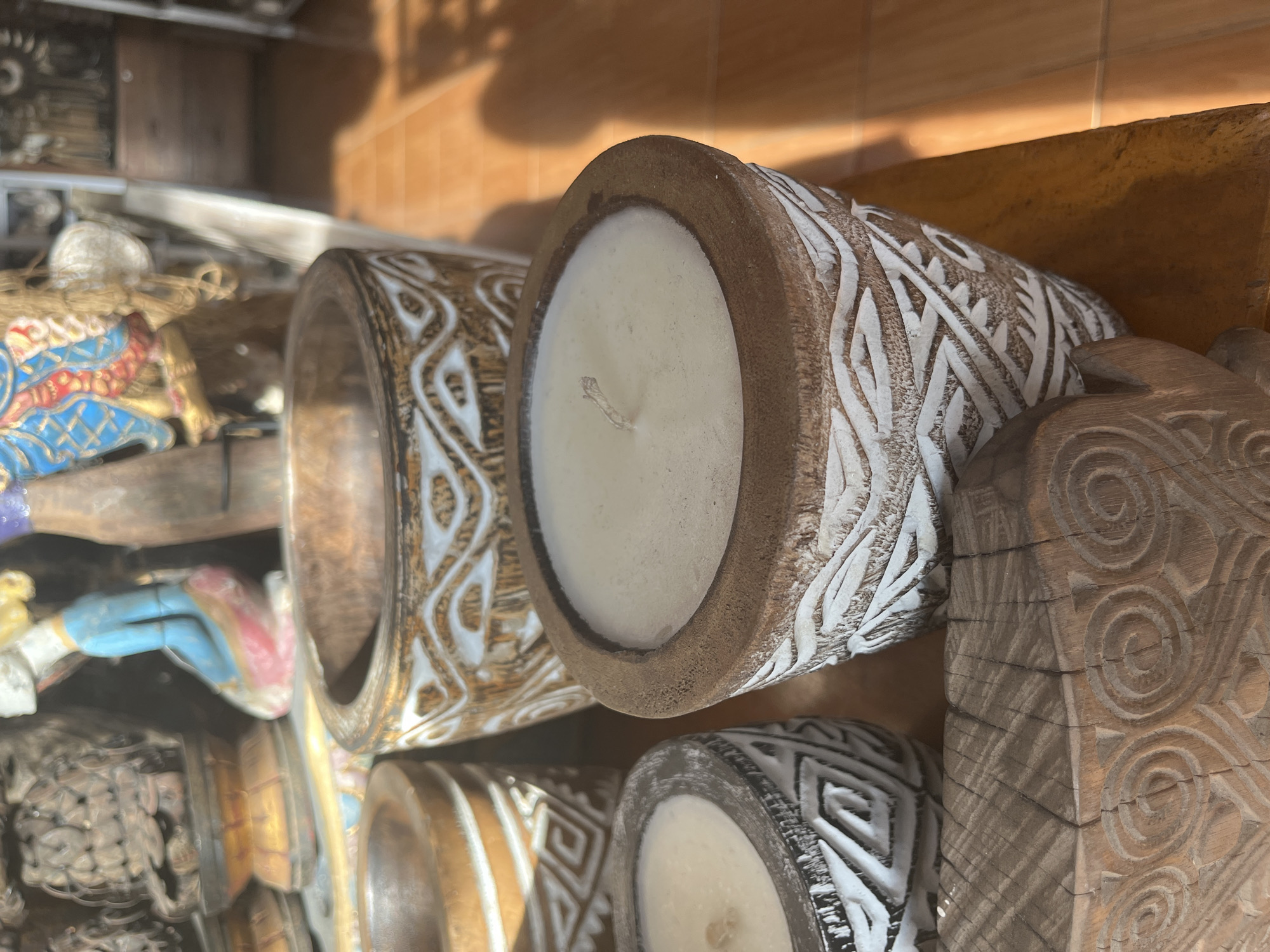 The image depicts several carved wooden objects, including candle holders with white wax fillings, on a shelf. The foreground features candle holders with intricate white carvings on a brown base. Adjacent to these are other carved objects, including a rectangular piece with spiral carvings. In the background, partially obscured, are additional figurines and wooden items, suggesting a display or storage area. The setting is likely a shop or display within the Ubud District, Indonesia, given the prevalence of traditional Balinese wood carvings.