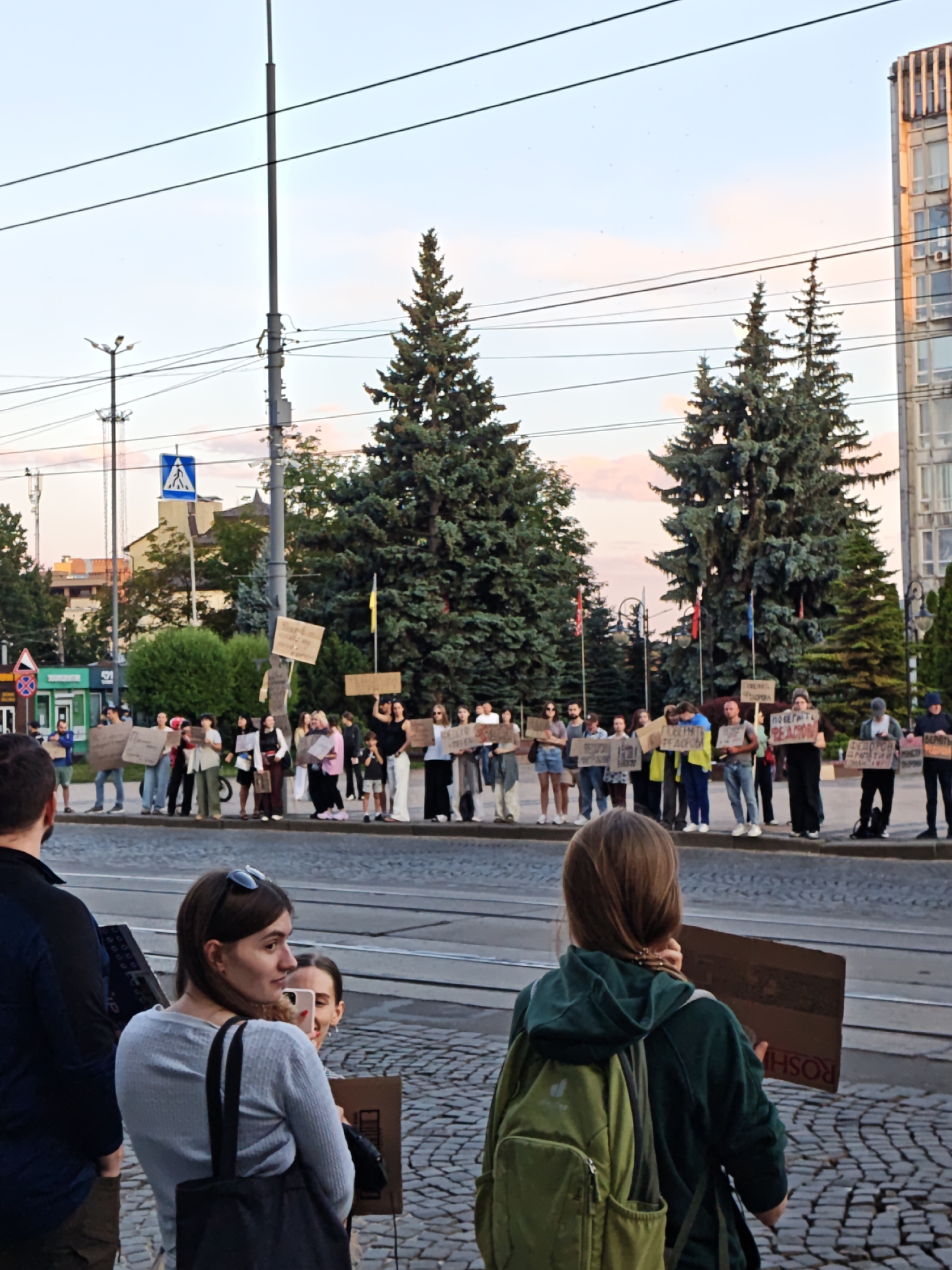 A crowd of people are holding signs at a street corner in Vinnytsia, Ukraine. The people are standing on a sidewalk next to a street with tram tracks. There are trees and a tall building in the background. The sky is a light blue with some clouds. The people are holding various signs, some of which appear to be in Ukrainian. The image appears to have been taken during the day.