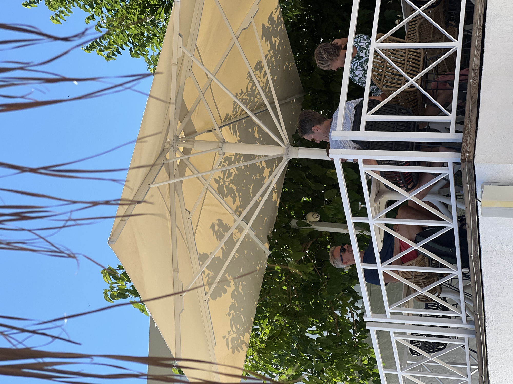 Three individuals are seated at a table on a white-railed balcony, under a large, cream-colored umbrella. The scene appears to be outdoors in a location with lush green foliage. The sky is a clear blue. The person on the far left is a man with grey hair, wearing sunglasses and a dark t-shirt with red shorts. He is looking to his left. The person in the middle is a man with grey hair, wearing a striped t-shirt and dark shorts, facing towards the right. The person on the far right is a woman with blonde hair, wearing a green and white patterned top, her back is partially turned towards the camera as she looks to her right. There are wicker chairs and tables on the balcony. A security camera is visible mounted on a pole behind the people. The overall setting suggests a restaurant or cafe with an outdoor seating area in Achladias, Greece.