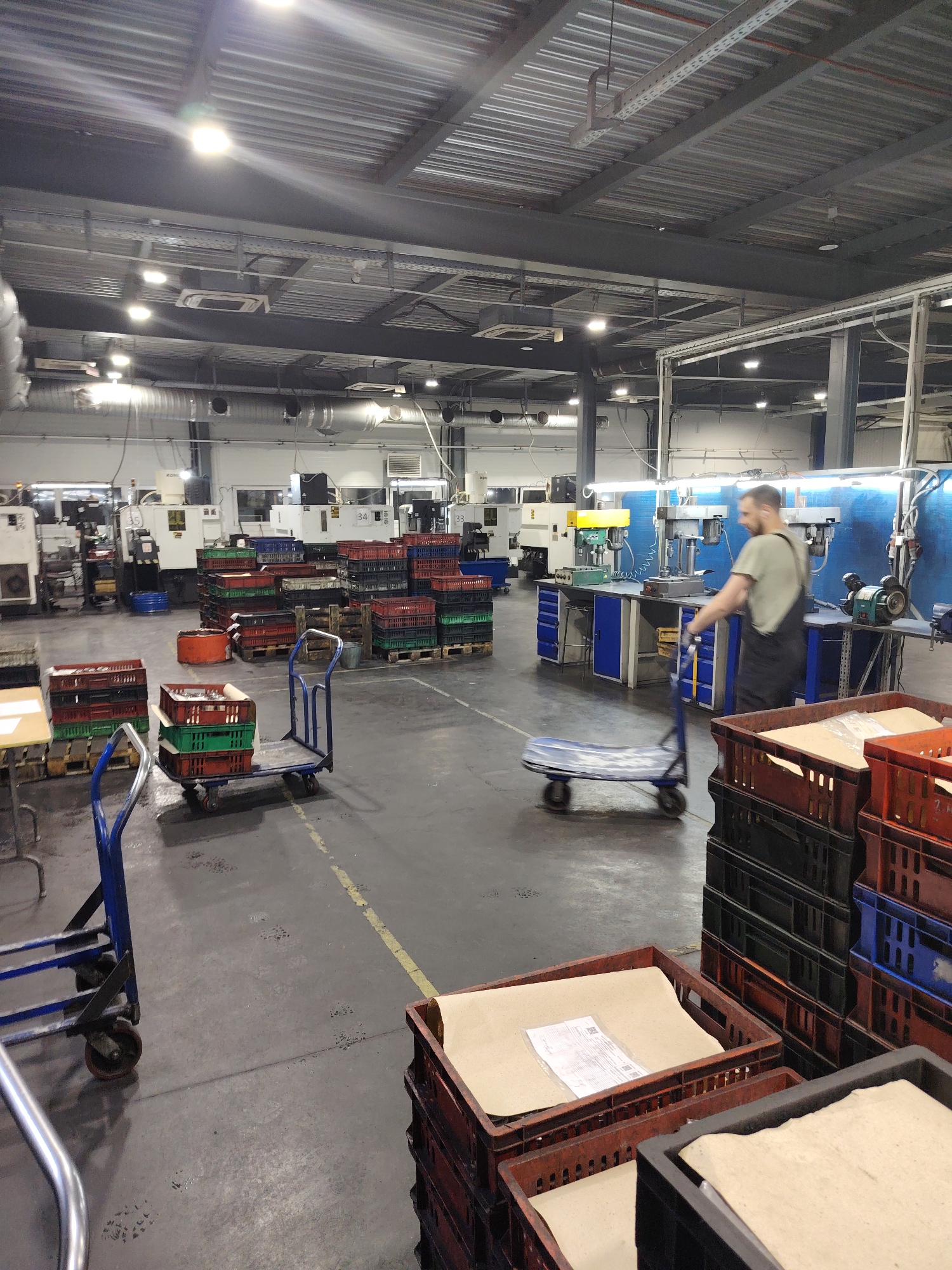 The image depicts the interior of a large, well-lit industrial facility, likely a manufacturing or processing plant. The location in the city of Noviy, Russia is not verifiable from the image.
The scene includes a man wearing work overalls and a light-colored t-shirt, using a wheeled platform trolley to move objects through the space. In the background, there are rows of industrial machines and workstations.
Several stacks of plastic crates, some with paper or other material inside, are visible throughout the area. Some crates are empty or contain only other plastic crates. Numerous metal support beams and ventilation ducts are visible overhead.
The floor is concrete and appears to be slightly worn or stained in places. Yellow lines are painted on the floor, presumably to mark pathways or designated areas. Several blue utility carts are also scattered around the space.