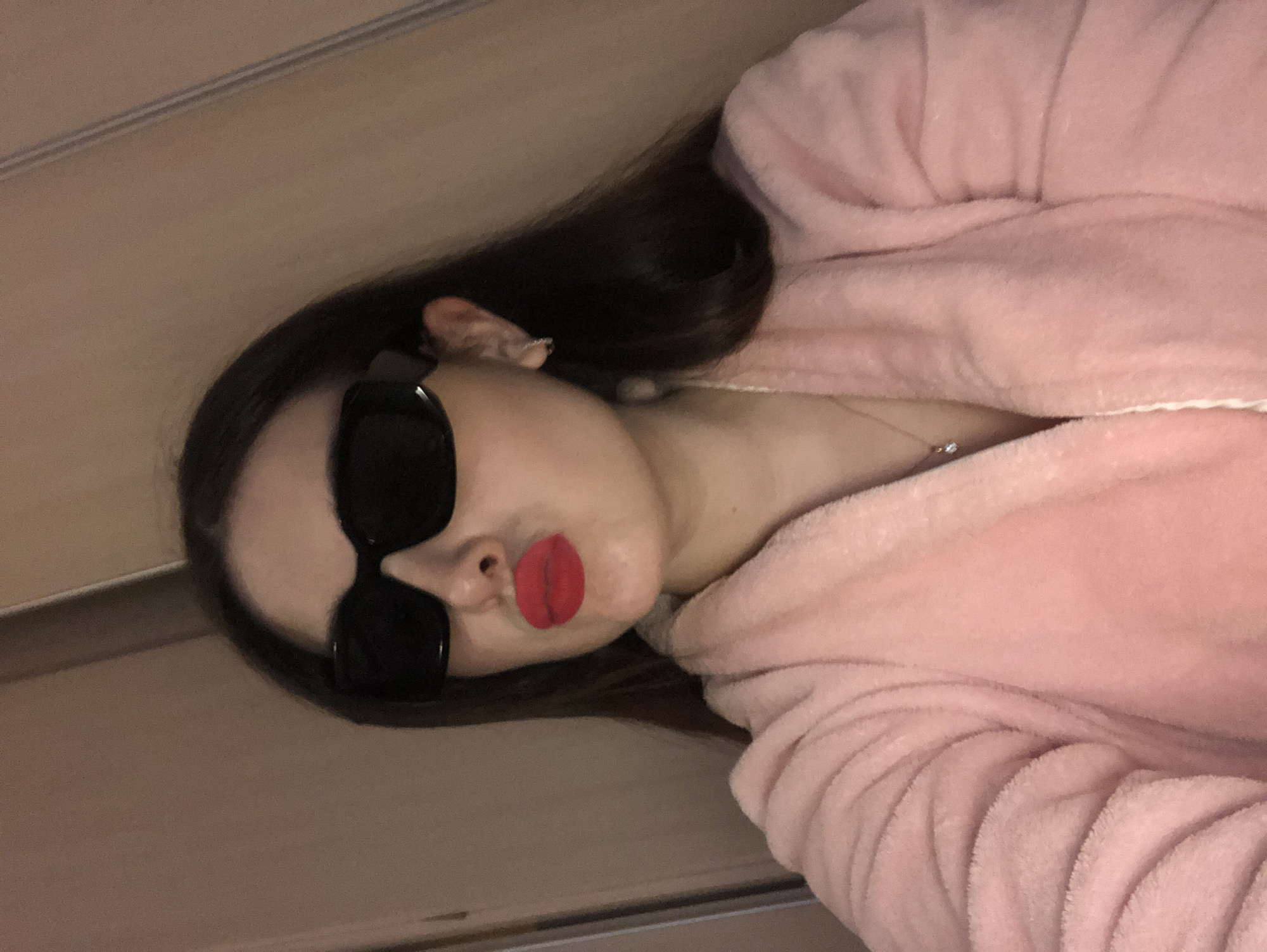 This media file features a close-up, tilted selfie of a person, likely female, from the chest up. The individual has long, dark hair and is wearing a soft, plush pink bathrobe. She is sporting large, black rectangular sunglasses and vivid red lipstick, with her lips pursed in a "kissing" or "duck face" expression. A delicate silver chain necklace with a small, clear round pendant is visible against her skin. The background consists of a plain, light brown or beige wall, with a faint horizontal line or trim running across it. The lighting is soft and appears to be artificial, illuminating the subject's face. The overall impression is one of a playful or sassy mood. No visible text is present in the image. While the location context of Noviy, Russia, was provided, there are no specific visual cues within the image to confirm this.