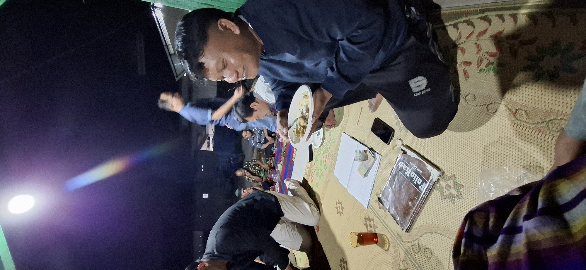 In Wonorejo, Indonesia, at night, a group of men are gathered on a woven mat outdoors, possibly for a meal. A bright overhead light casts a lens flare and a rainbow effect across the dark sky. One man in the foreground, wearing a dark long-sleeved shirt and black pants, is smiling and eating from a white plate held in his hands. Several other men are seated or kneeling around him on the mat. To the left, a man in a black shirt and white pants is kneeling. In the background, other men are visible in the dim light. On the mat in front of the foreground man are a glass of amber liquid, an open notebook with papers and money, a smartphone, and a book with the title "FolioKas" visible. The environment appears to be an outdoor residential area, with hints of buildings and possibly a green tarp in the background.