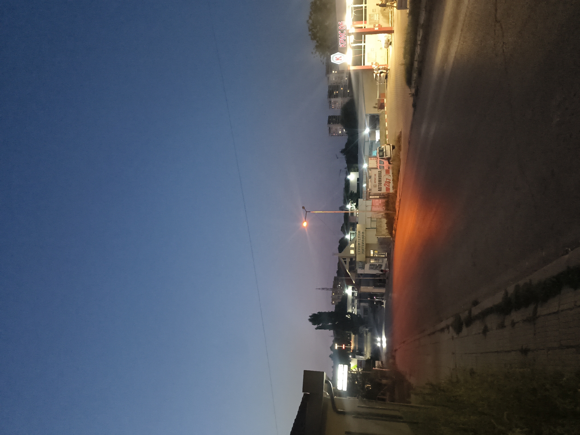 The image depicts a street scene in Shumen, Bulgaria, during dusk or early evening. A street with a sidewalk is visible in the foreground, with a paved road extending into the mid-ground. On the right side of the road, there is a car wash facility with illuminated signage and several cars present. One car is being attended to by two individuals, while another person walks away from the facility. Across the street, on the left side, there are several buildings, including one with illuminated signs and what appears to be a commercial establishment. Further in the distance, there are apartment buildings with lights on. The sky is a deep blue, transitioning to a lighter shade near the horizon, with the silhouette of trees and a radio tower visible against the sky. Streetlights are illuminated, casting a warm glow on the road. The overall impression is of a commercial area at the edge of a residential zone.