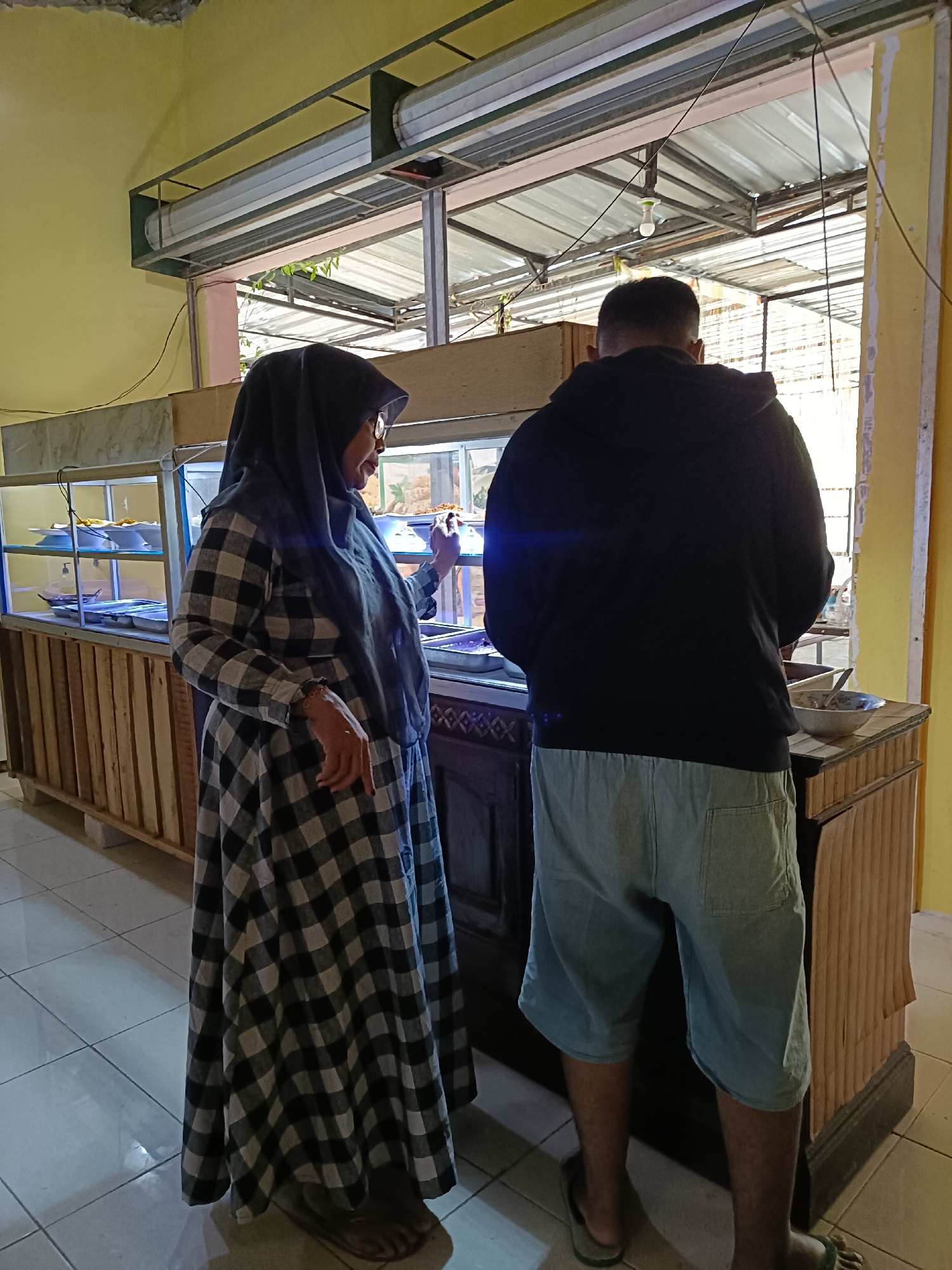 The image shows a woman in a black and white checkered dress and a black hijab standing in front of a food display case. A man wearing a black hoodie and light blue shorts stands to her right, also facing the food display. The food display case has wooden framing and contains various food items in trays. Behind the display case is a window with a view of what appears to be an outdoor area with a corrugated metal roof supported by metal beams. The floor is tiled with light-colored square tiles. The wall behind the woman is painted yellow. The scene is likely a food stall or a small eatery in Pordapor, Indonesia. The woman appears to be selecting food, while the man is looking in the same direction.