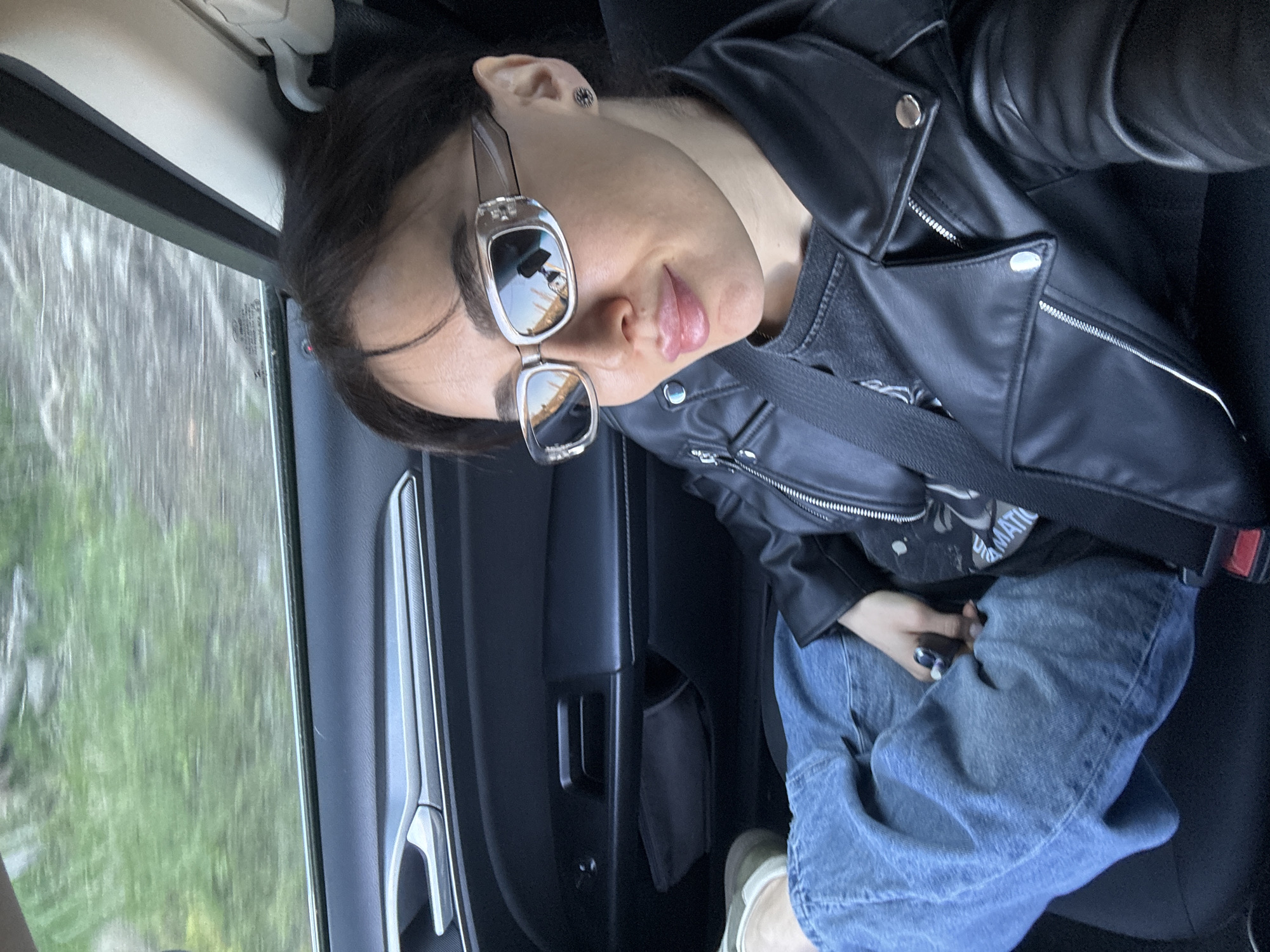 A woman wearing sunglasses and a black leather jacket sits in a car. She is also wearing blue jeans. The interior of the car is dark, with a silver door handle visible. The window shows green foliage blurring by, suggesting the car is moving. The woman's face is angled toward the camera. Based on the registration code on the sunglass frames, it is believed that the photo was taken in Tbilisi, Georgia.