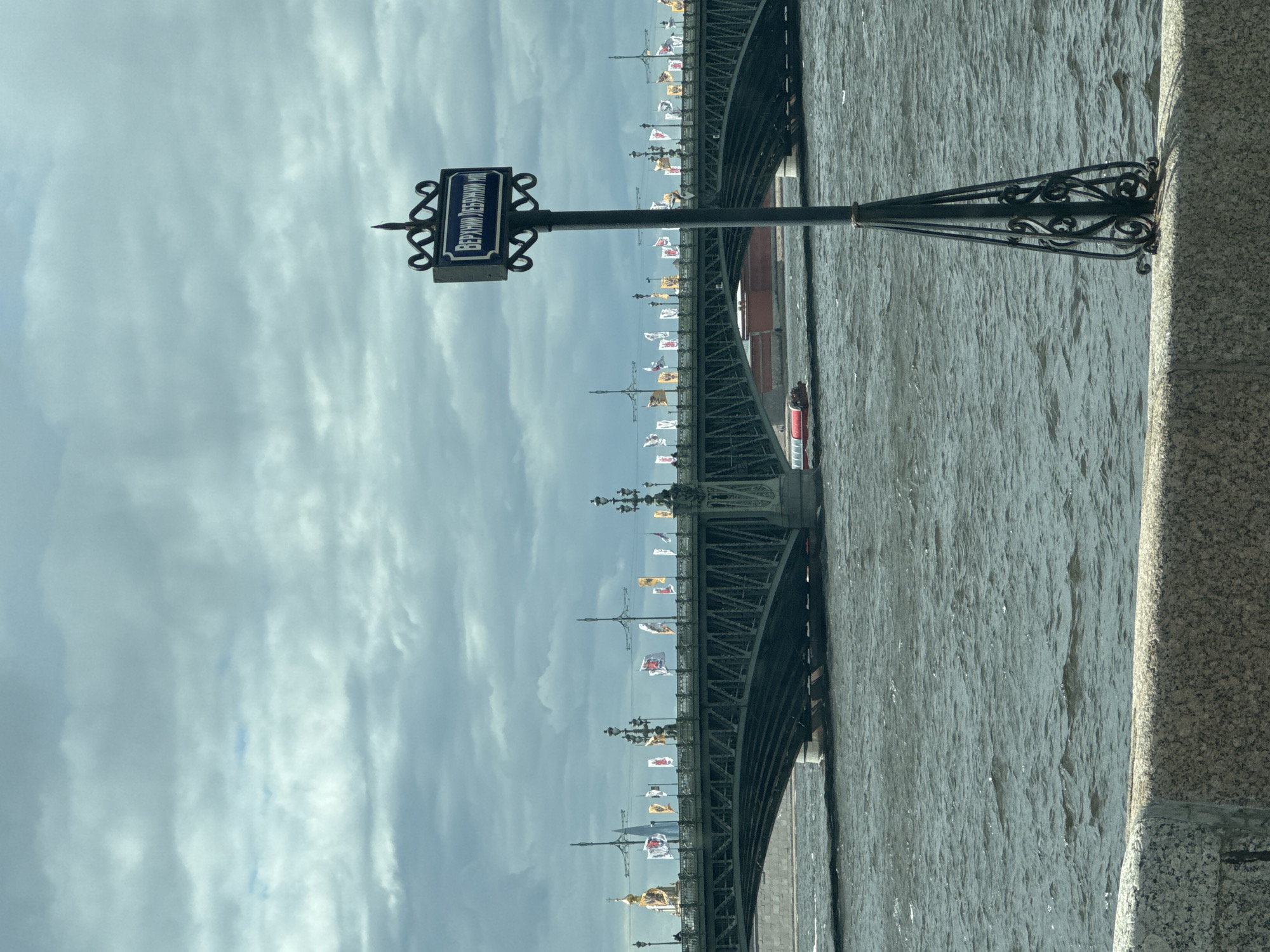 The image shows the Liteyny Bridge in Saint Petersburg, Russia, as seen from the embankment. The bridge is decorated with flags, and the Neva River flows beneath it. The sky is cloudy. A street sign reading "Верхний Лебяжий мост" (Upper Swan Bridge) is visible.