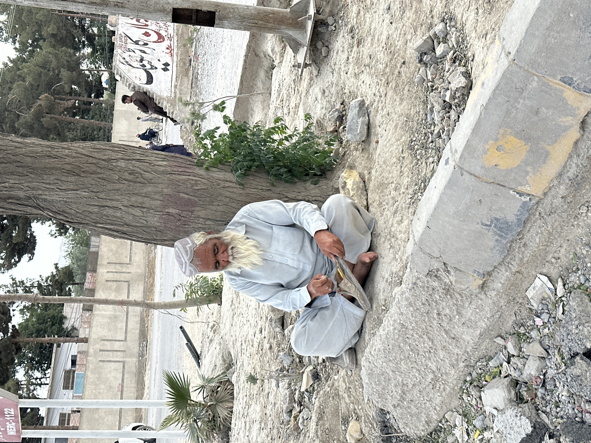 An elderly man with a long white beard and a white cap sits on the ground in Quetta City Tehsil, Pakistan. He wears a light blue long-sleeved shirt and light grey pants. He's mending a shoe. The ground is dirt, concrete fragments, and small stones. A curb runs along the right side. In the background, there are trees, buildings, and other people walking along the street. A sign post with the words "MERC-1122" is in the lower left corner.