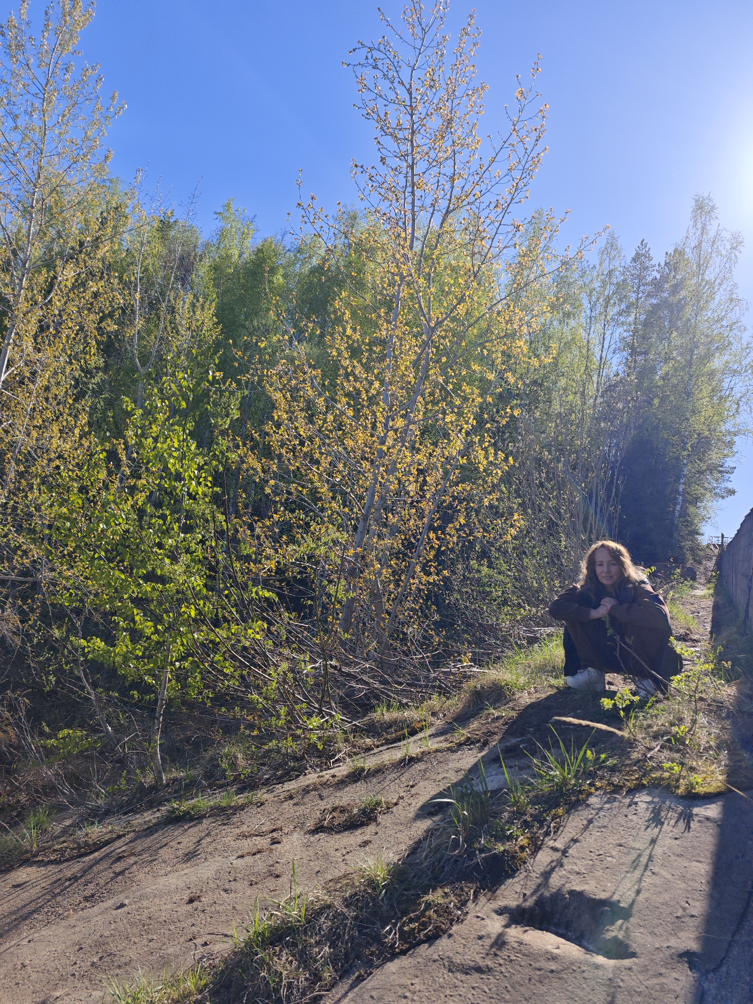 The photograph was taken in an outdoor environment, possibly near or in the city of Chernushka, Russia. The setting appears to be a sloped, somewhat rocky or earthen area with vegetation that includes trees and grass. A woman with red hair is squatting on the slope, looking towards the camera. She is wearing a dark, possibly brown, jacket or coat. The background consists of various trees, some with new foliage emerging, indicating a springtime scene. The sky is a clear blue. The light is bright, suggesting it's daytime, with the sun casting shadows across the ground. The foreground includes some rocks and patches of grass.