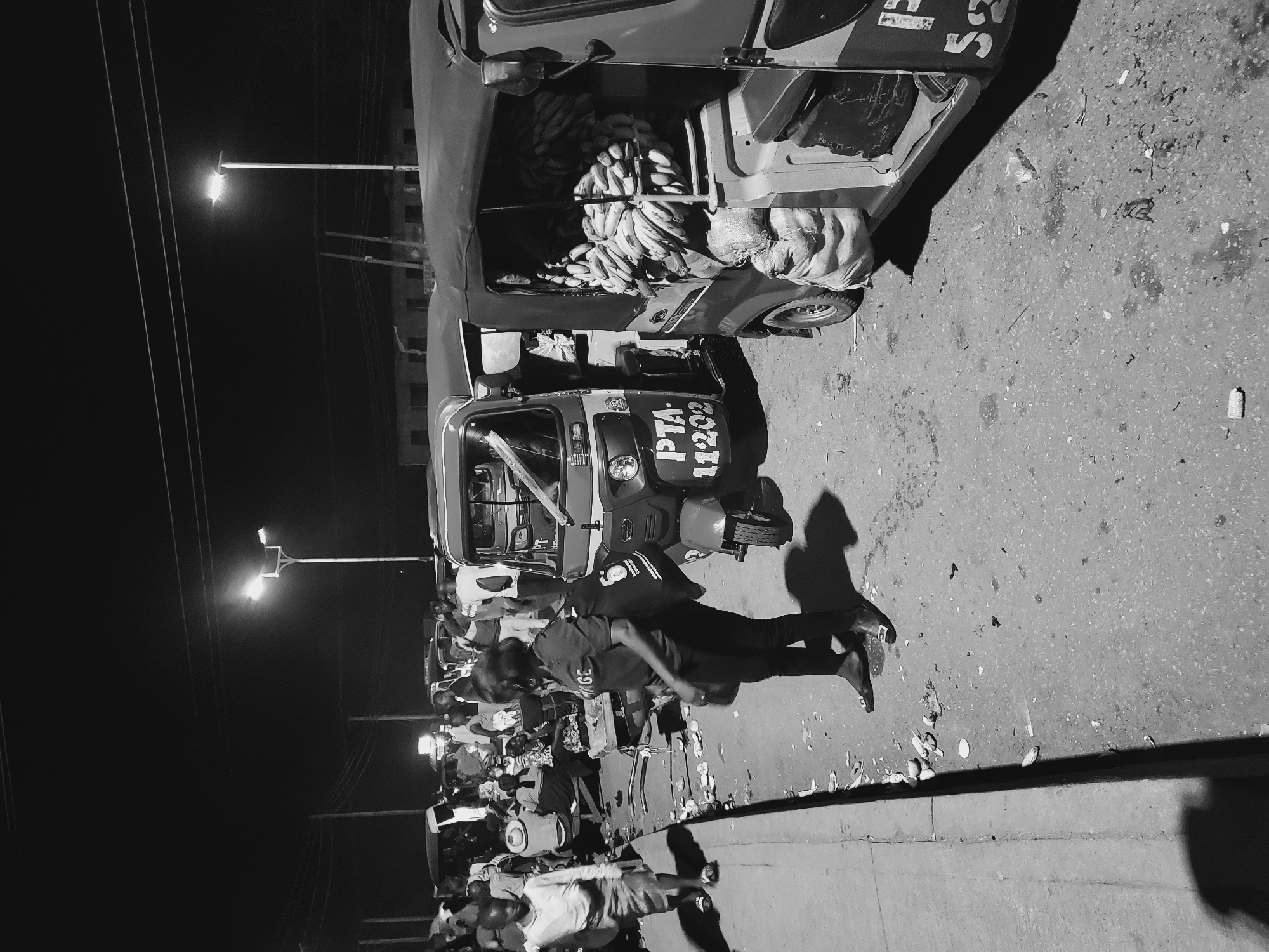 This black and white photograph captures a bustling street scene at night in Bukuru, Nigeria. The scene is illuminated by several streetlights and vehicle lights, casting long shadows across the ground.
In the foreground, a prominent auto-rickshaw (tuk-tuk) is visible, heavily loaded with numerous bunches of bananas and what appear to be sacks, suggesting it's transporting goods. The vehicle has text "PTA-11202" clearly visible on its front. Another auto-rickshaw is partially visible next to it, also appearing to carry goods, with indistinct text on its side.
Numerous people are gathered along the street, indicative of a busy marketplace or commercial area. One person, dressed in dark clothing with "IGE" visible on their shirt, is walking in the mid-ground, casting a distinct shadow to their left. Others are congregated further back, some standing near other parked vehicles or stalls.
The ground is a paved surface, likely a road, with a concrete curb running along the bottom edge, separating it from a pedestrian area or shoulder. Scattered debris and litter are visible on the ground, possibly fruit peels from the market activity. Power lines stretch across the dark night sky. The overall atmosphere suggests a lively night market or a commercial hub active after dark.