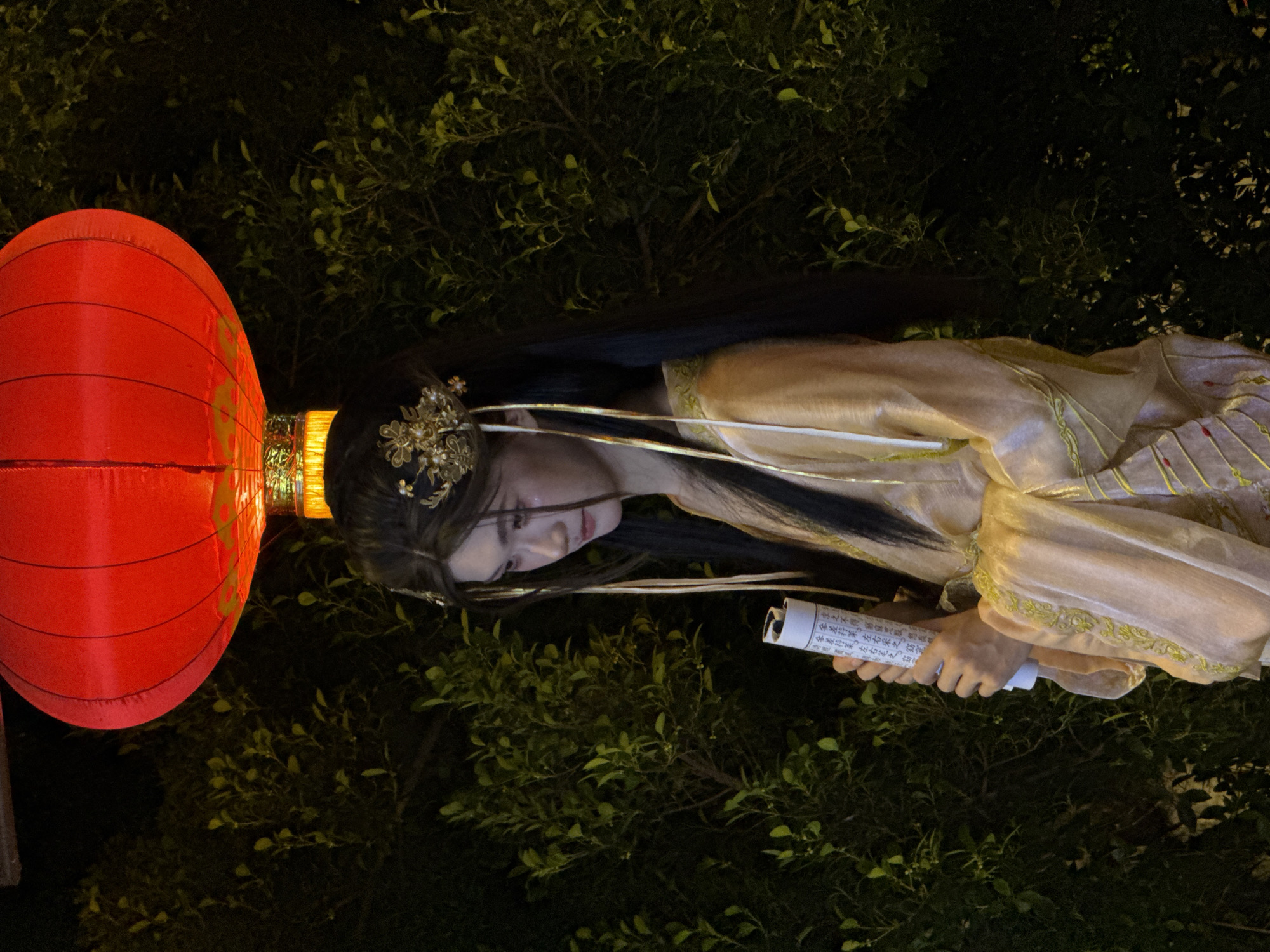 A young woman in traditional Chinese attire stands outdoors at night, illuminated by a red lantern hanging above. She wears a pale gold, flowing robe with intricate embroidery and has her dark hair styled with ornate gold hairpins and dangling ribbons. The woman is looking down and holding a rolled scroll tightly in her hands. She is positioned against a dark backdrop of dense green foliage, suggesting an outdoor setting possibly within a garden or park in the Yuzhong District of China. The overall atmosphere is serene and evocative of ancient Chinese culture.