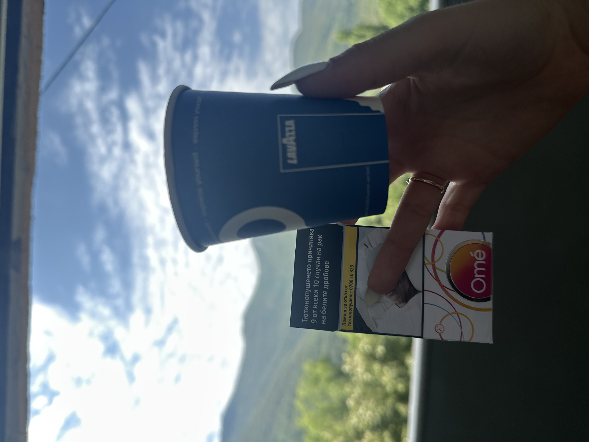 A person's hand, adorned with a delicate ring and long, white fingernails, holds a blue paper coffee cup and a pack of cigarettes with a warning label in Bulgarian against a backdrop of mountains and sky. The cup has the Lavazza logo. The cigarette pack displays a warning about smoking causing 9 out of 10 cases of lung cancer, along with a helpline number. The scene is an outdoor setting, likely on a balcony or terrace with a view of lush green mountains and a cloudy blue sky, suggestive of a location like Vratsa, Bulgaria.