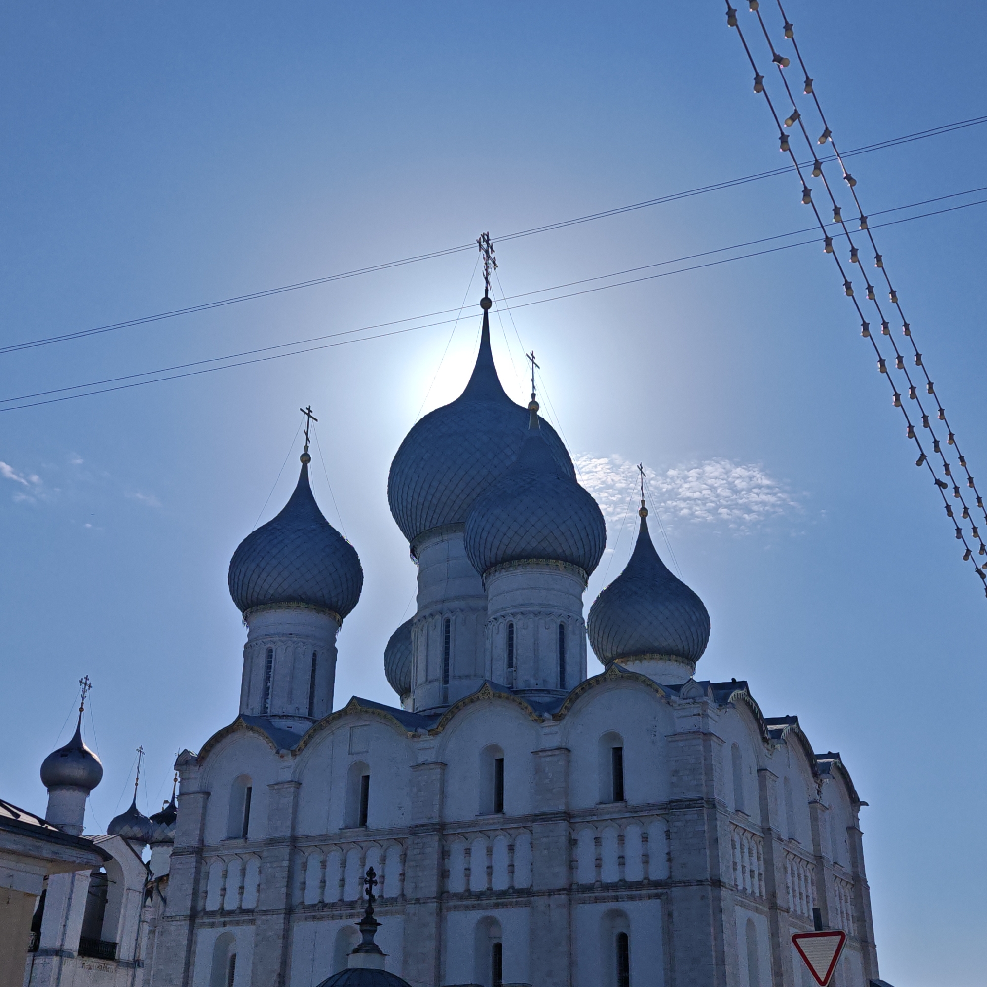 The image shows a low-angle view of a white stone cathedral with multiple blue-gray domes topped with crosses. The sun is directly behind the central dome, causing a strong backlight effect. Two sets of power lines cross the upper portion of the frame, and a string of lights hangs diagonally in the upper right corner. In the lower right, there's a road sign with a white triangle on a red background. Part of another building is visible on the left side. The photo was taken in Ново-Никольское, Russia.