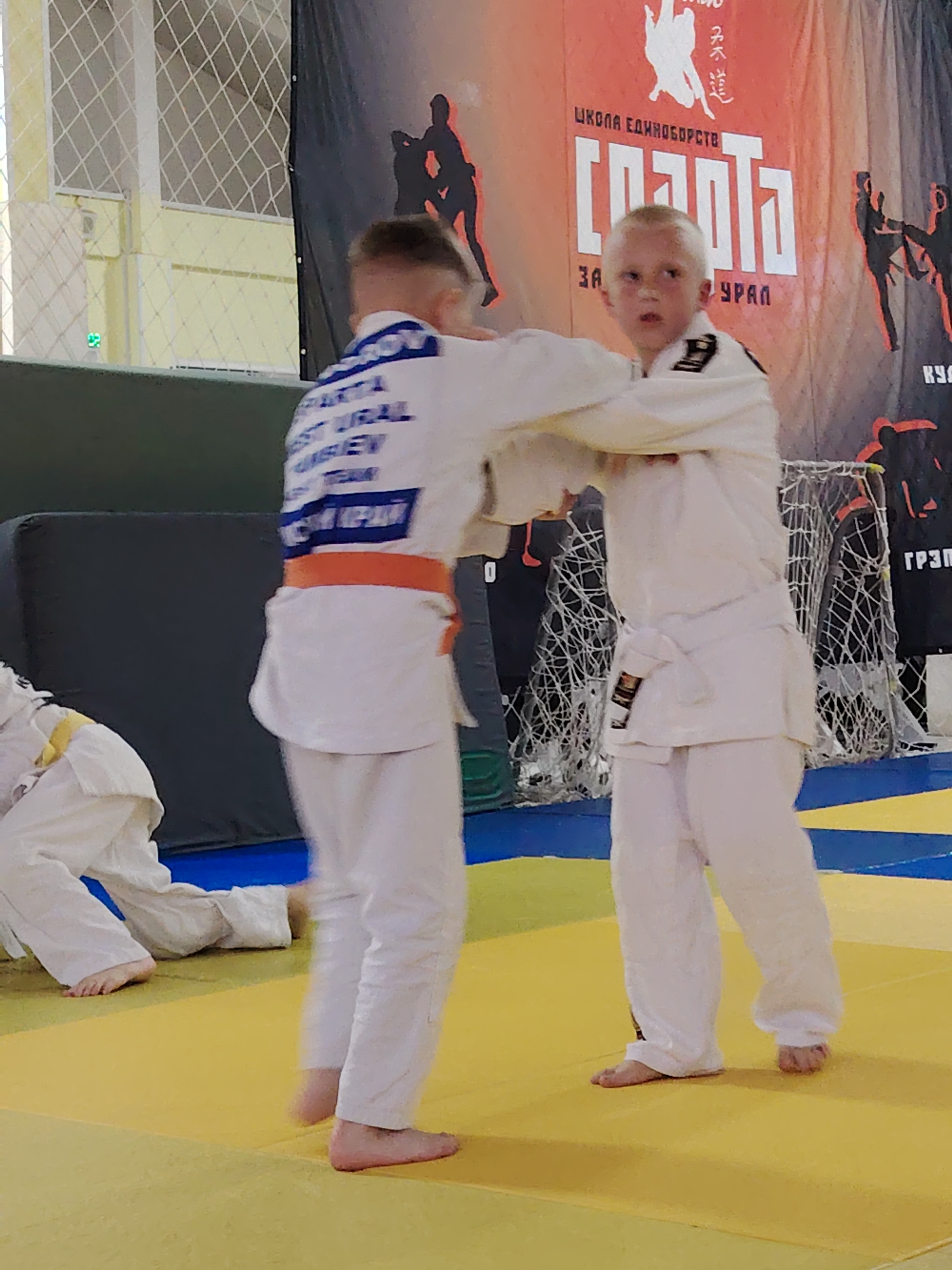 Two young boys in white judo uniforms are engaged in a sparring match on a yellow and blue mat. The boy on the left has an orange belt and blue text on the back of his uniform. They are barefoot. In the background, there is a banner with Russian text and silhouettes of martial artists. The banner indicates the location is the city of Chaikovsky, Russia. There is a net-like barrier and a dark gray rectangular object to the left. Another child in a white judo uniform is partially visible on the left side of the frame.