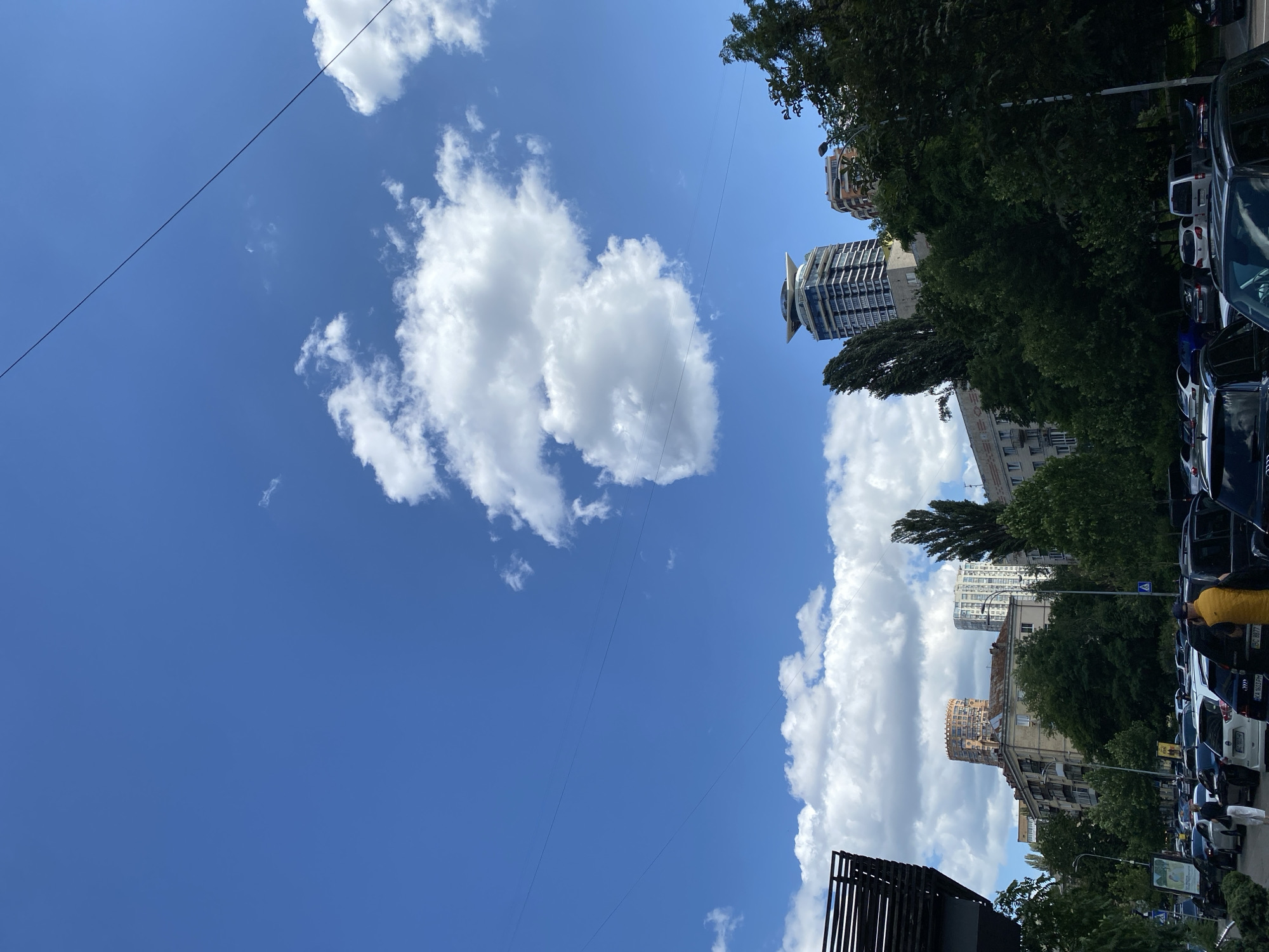 This image captures a street view in Kyiv, Ukraine, on a clear day with scattered white clouds. The scene is dominated by a bright blue sky. In the foreground, several parked cars line a street, with people visible walking and standing near them. A distinct, round, tower-like building with a crenellated top is visible in the mid-ground, featuring a reddish-brown exterior. To its right, a modern, sleek skyscraper with a distinctive winged roof structure stands out. Lush green trees partially obscure some of the buildings. Power lines stretch across the sky, indicating urban infrastructure. The overall impression is a typical urban landscape with a mix of older and contemporary architecture under a vast sky.
