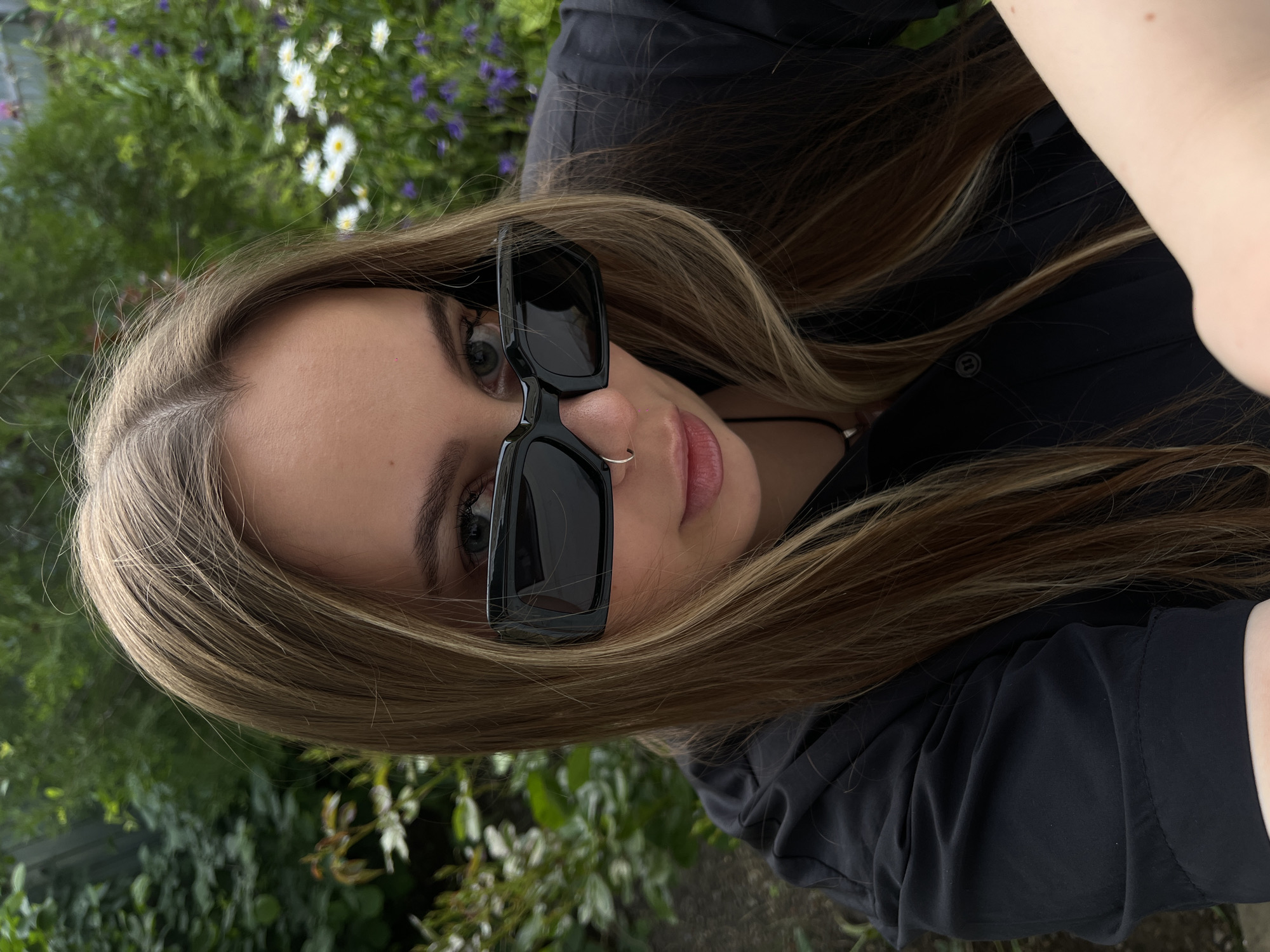 A close-up, tilted portrait features a person, likely female, with long, light brown hair framing her face. She is looking directly at the camera. Her head is positioned on the left side of the frame due to the image's rotation.
She wears large, rectangular black sunglasses that cover the upper part of her face. A small silver hoop piercing is visible on her right nostril. Her left eye (viewer's right) is visible above the sunglasses, appearing to be blue or grey, and her lips are slightly parted. She is dressed in a dark, possibly black, long-sleeved top or jacket, visible on the right side and bottom of the frame.
The background is an out-of-focus outdoor setting, filled with lush green foliage and scattered small white and purple flowers, suggesting a garden or natural area. The lighting is natural, indicating it is daytime. The overall impression is a casual, direct gaze set against a soft, verdant backdrop. No text is visible in the image.