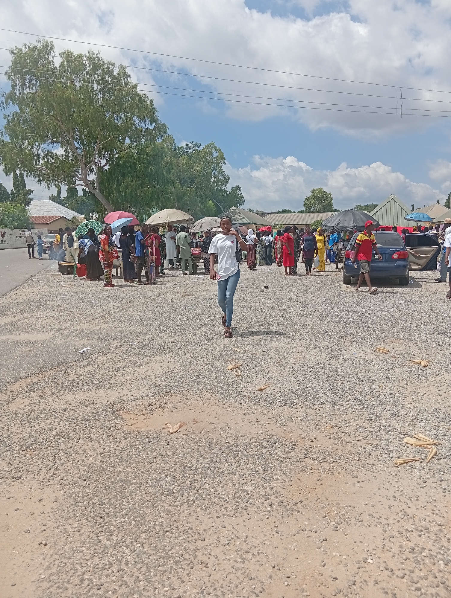 The image depicts an outdoor scene, likely a market or a gathering, on a paved surface covered with gravel. In the foreground, a young woman with dark hair walks towards the camera, wearing a white t-shirt and blue jeans. Behind her, a blue sedan is parked with its passenger door open. Scattered throughout the gravel are dried plant stalks, possibly corn husks.
In the mid-ground, a crowd of people are visible, many standing under umbrellas, suggesting a sunny day. Several individuals are wearing colorful traditional clothing. There are also market stalls or vendors visible, with goods displayed on tables and carts. A few buildings and trees are present in the background.
The overall impression is of a bustling outdoor event. The presence of a specific sign visible on a building in the background, "TARABA STATE SECRETARIAT," indicates that the location is within Jalingo, the capital city of Taraba State in Nigeria. The environment appears to be a public area, possibly a roadside market adjacent to administrative buildings. The weather seems to be clear with some clouds in the sky.