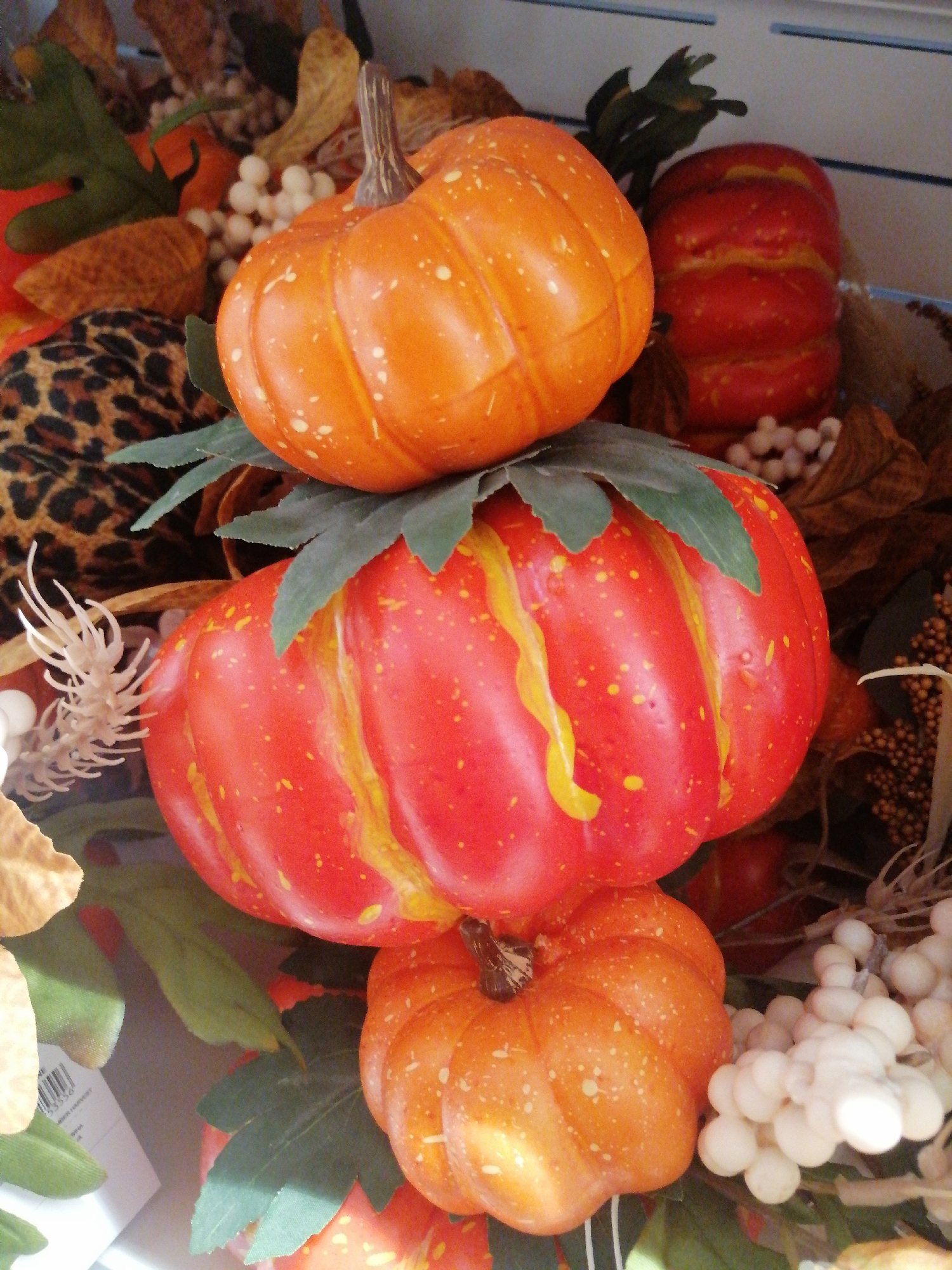 An arrangement of artificial pumpkins and fall foliage is displayed. A small, orange pumpkin with white speckles is positioned at the top. Below it, a larger, red and yellow striped pumpkin is centrally located. At the bottom, another small, orange pumpkin with white speckles is visible. The arrangement includes green leaves, dried leaves, white berries, and a piece of leopard-print fabric. The background is out of focus, but appears to be a shelf or display surface. There are no people present, and no actions are taking place. The scene is static. The image does not provide any information about its location in the city of Kremenchuk, Ukraine.