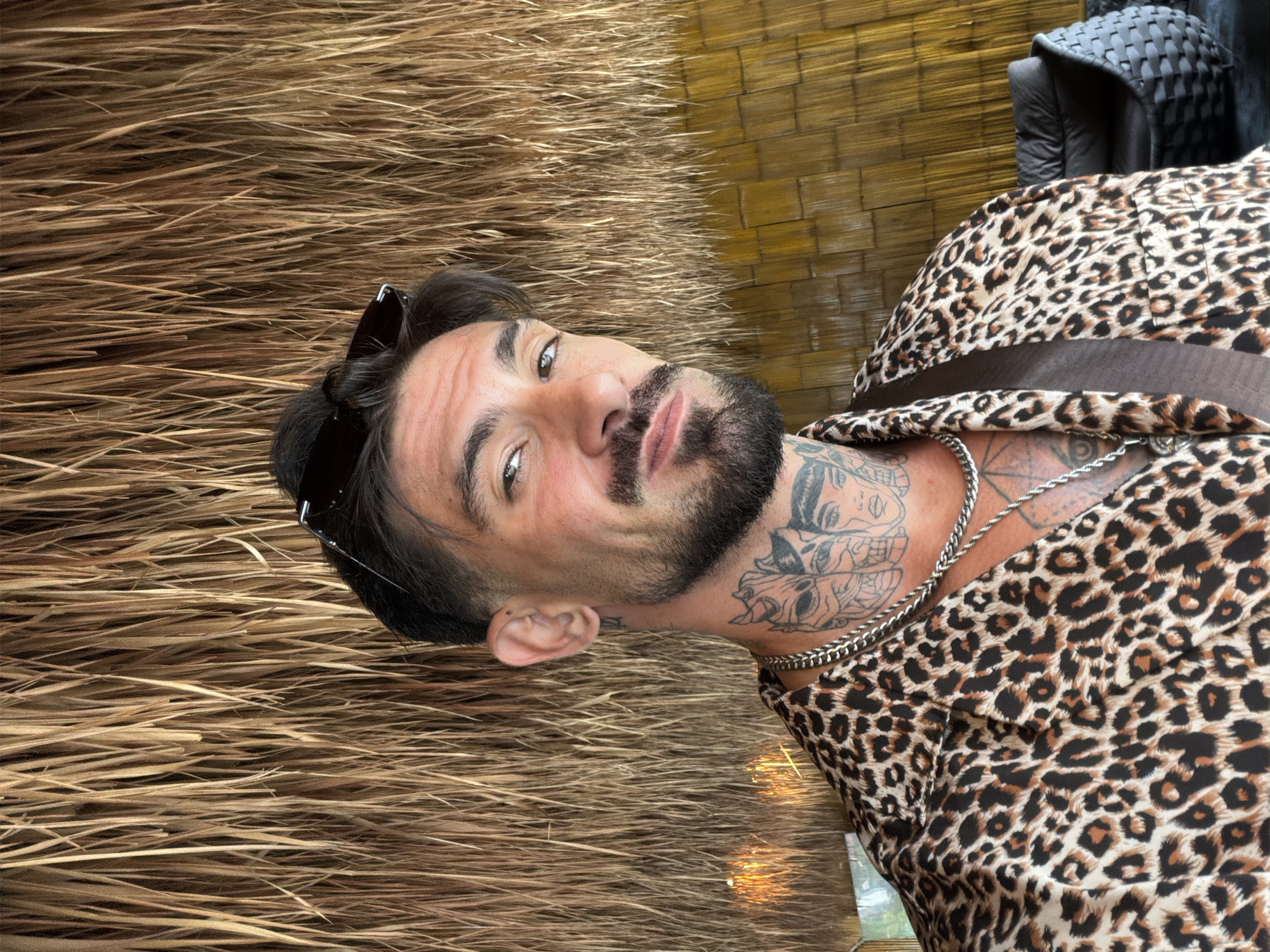 A man with a dark beard and neck tattoos is positioned against a background of thatch and bamboo. He wears a leopard-print shirt, a silver chain, and sunglasses pushed up on his head. The photo was taken in Ban Khlong Khian บ้านคลองเคียน, Thailand.