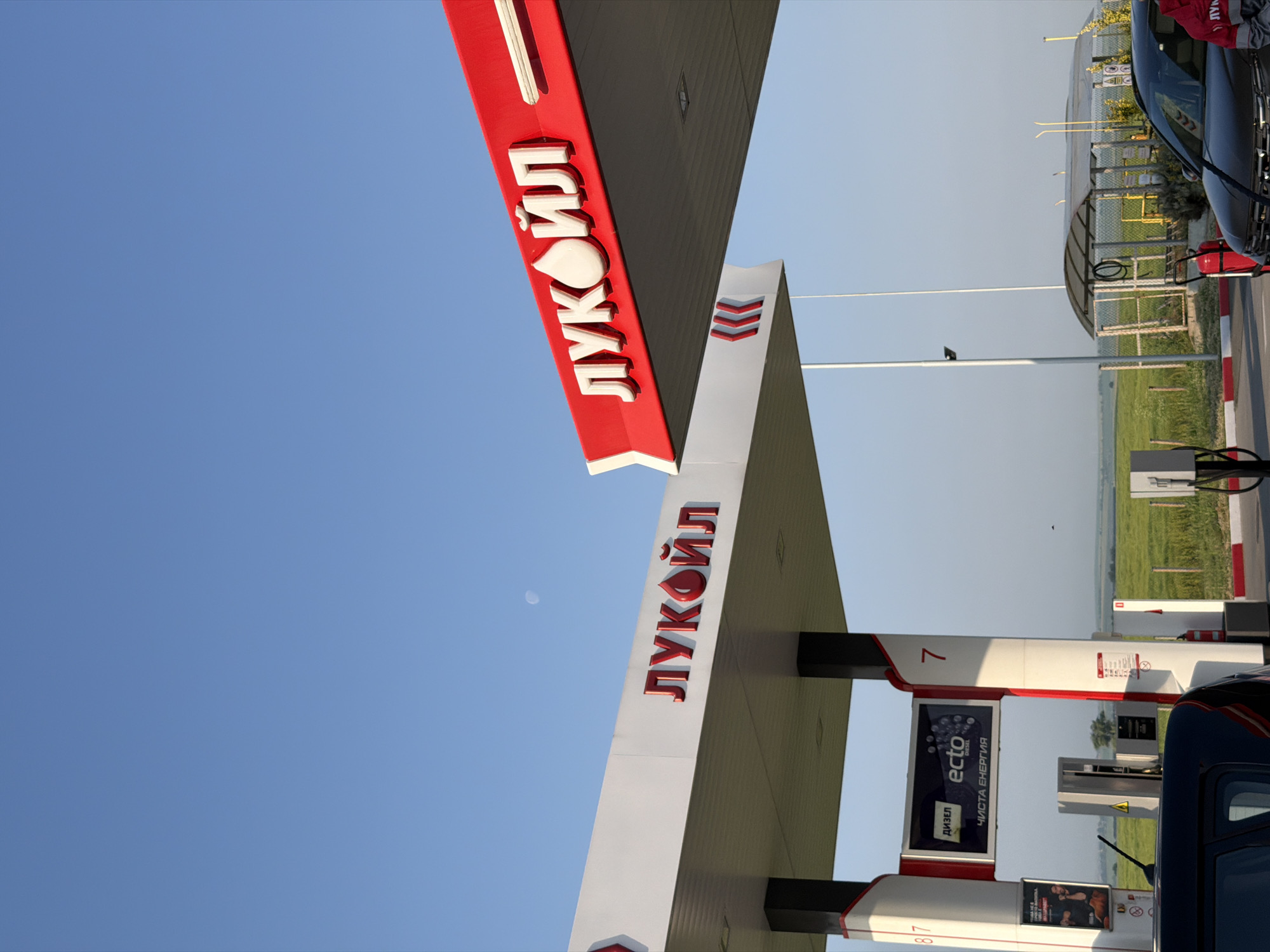 The image captures a vibrant, high-angle view of a Lukoil gas station under a clear, bright blue sky, presumably in Vodenichane, Bulgaria, given the Cyrillic script. The perspective is tilted, showing the underside and sides of two prominent red and white canopies that extend over the fueling area. Both canopies feature the distinct "ЛУКОЙЛ" (LUKOIL) logo in white, three-dimensional letters against the red stripe. The larger canopy also displays red chevron markings.
At the lower right, a black car is partially visible, parked next to gas pumps. One pump pillar clearly shows the number "7" and an advertisement for "ectо ДИЗЕЛ" (Ecto Diesel) with the tagline "ЧИСТА ЕНЕРГИЯ" (Clean Energy). Another pump, partially visible at the very bottom, shows the number "87". A red fire extinguisher is positioned near the car. The ground features red and white striped curbing, delineating the service area.
In the background, beyond the gas station structures, a simple, covered shelter (possibly a car wash or service bay) with a mesh fence is visible. Green agricultural fields stretch into the distance under the open sky, suggesting a semi-rural setting. A small, faint moon is discernible in the upper left portion of the sky, indicating it is daytime, likely morning or late afternoon. The overall weather appears sunny and calm, with no clouds. No people are explicitly visible, but their presence is implied by the active gas station environment.