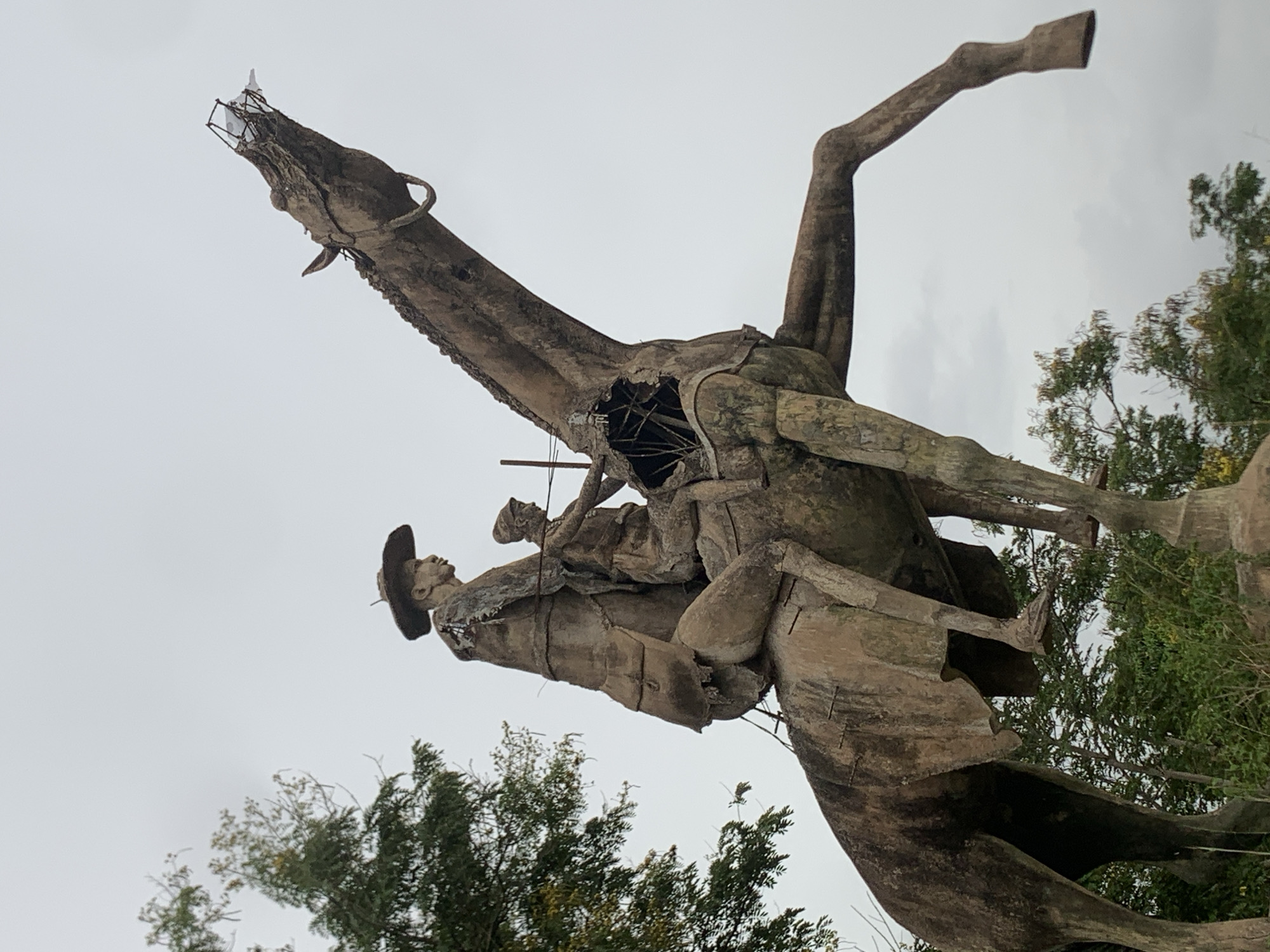The image depicts a deteriorated concrete sculpture of a cowboy riding a rearing horse. The sculpture exhibits significant damage, including a gaping hole in the horse's chest exposing the internal metal framework, and the front of the horse's head appears to be missing concrete or other material. The horse's tail and one of its legs are also absent, though the leg and hoof remain. The cowboy, dressed in a hat and coat, is seated atop the horse with another person behind him. The statue is located outdoors, under an overcast sky, and is surrounded by trees and vegetation. It is identified as being located in Gura Top, Nigeria.