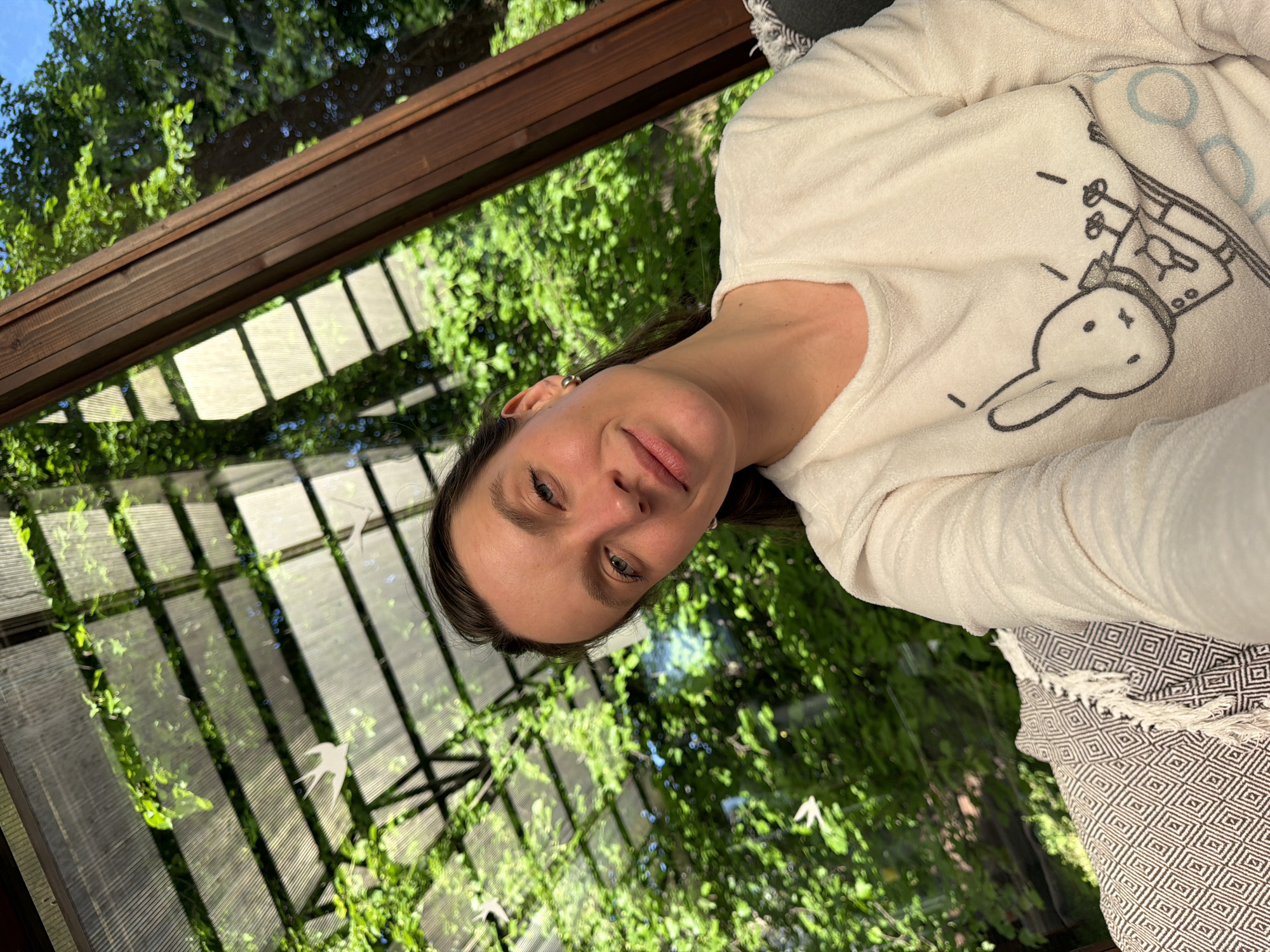A woman wearing a white, long-sleeved shirt with a cartoon bunny is sitting inside what appears to be a sunroom or greenhouse. The room has a glass or translucent roof and walls, with a wooden frame. Outside the room, there are green trees and vegetation visible through the glass. A patterned throw blanket is draped near the woman. The woman is smiling and looking at the camera. The location is Tarasovka, Russia.
