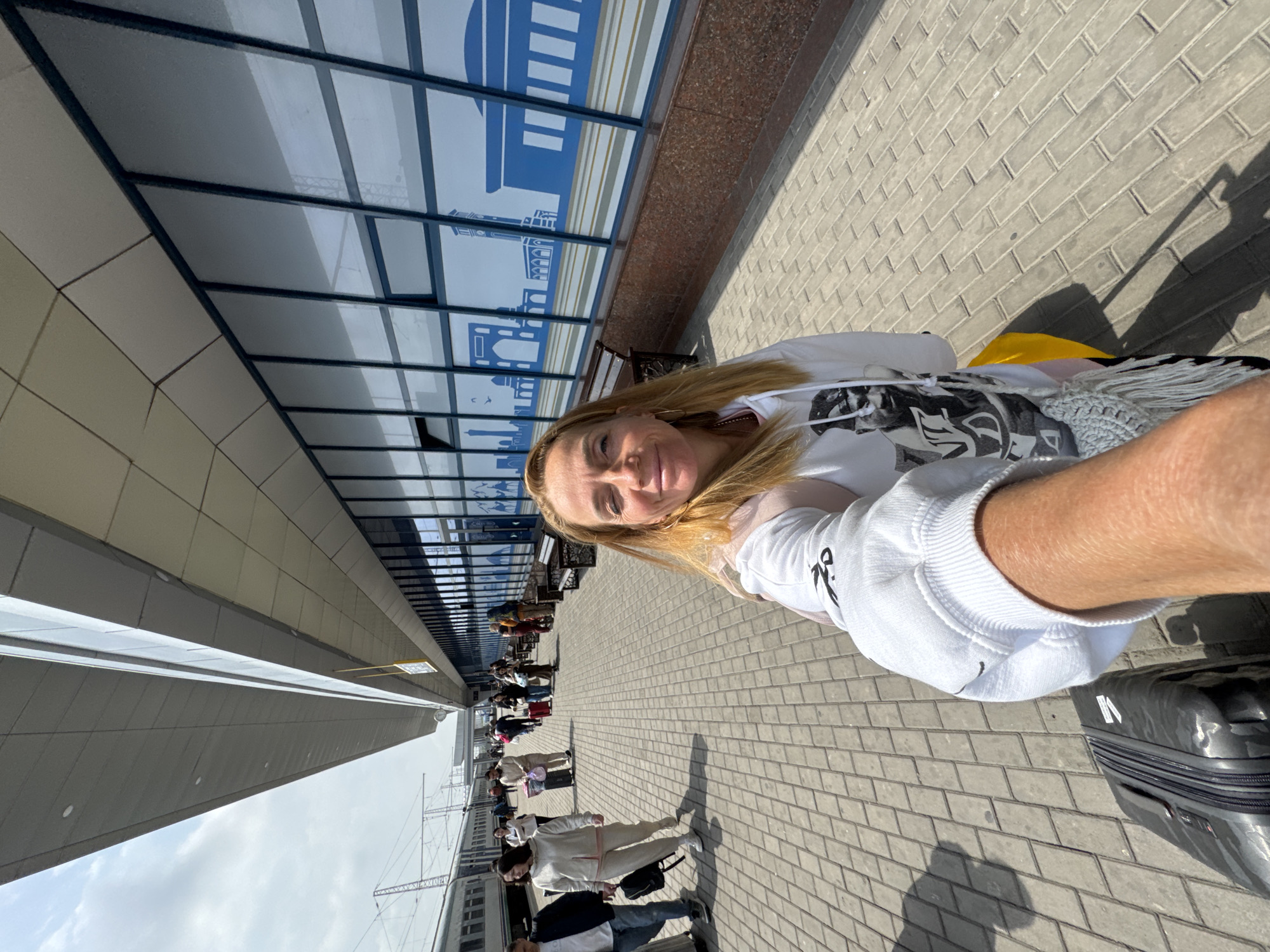 A selfie of a woman with a fair complexion and blonde hair standing outdoors on a brick-paved surface. She is wearing a white sweatshirt. Behind her is a building with a glass facade decorated with blue silhouettes of landmarks. Several people are seen in the background along the walkway, some pulling luggage. A train is visible in the far background. A suitcase is visible next to the woman. The location appears to be the Tashkent Railway Station in Tashkent, Uzbekistan.