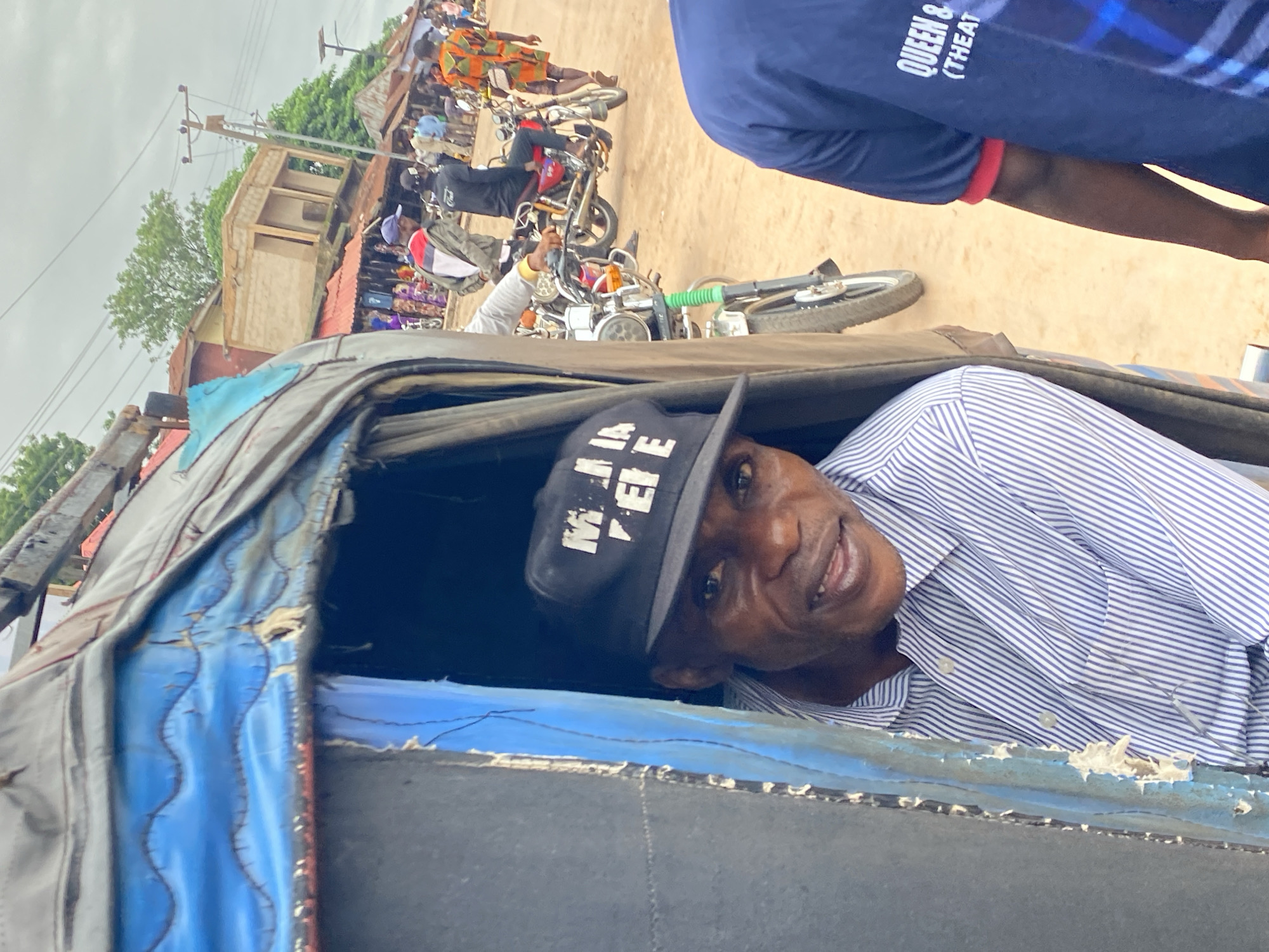 A man in a striped shirt and a black "Mania Pepe" cap is seen inside a blue tuk-tuk in Nguzu Edda, Nigeria. Outside, motorcycles and people are visible on a dusty road. A building under construction and power lines are in the background.