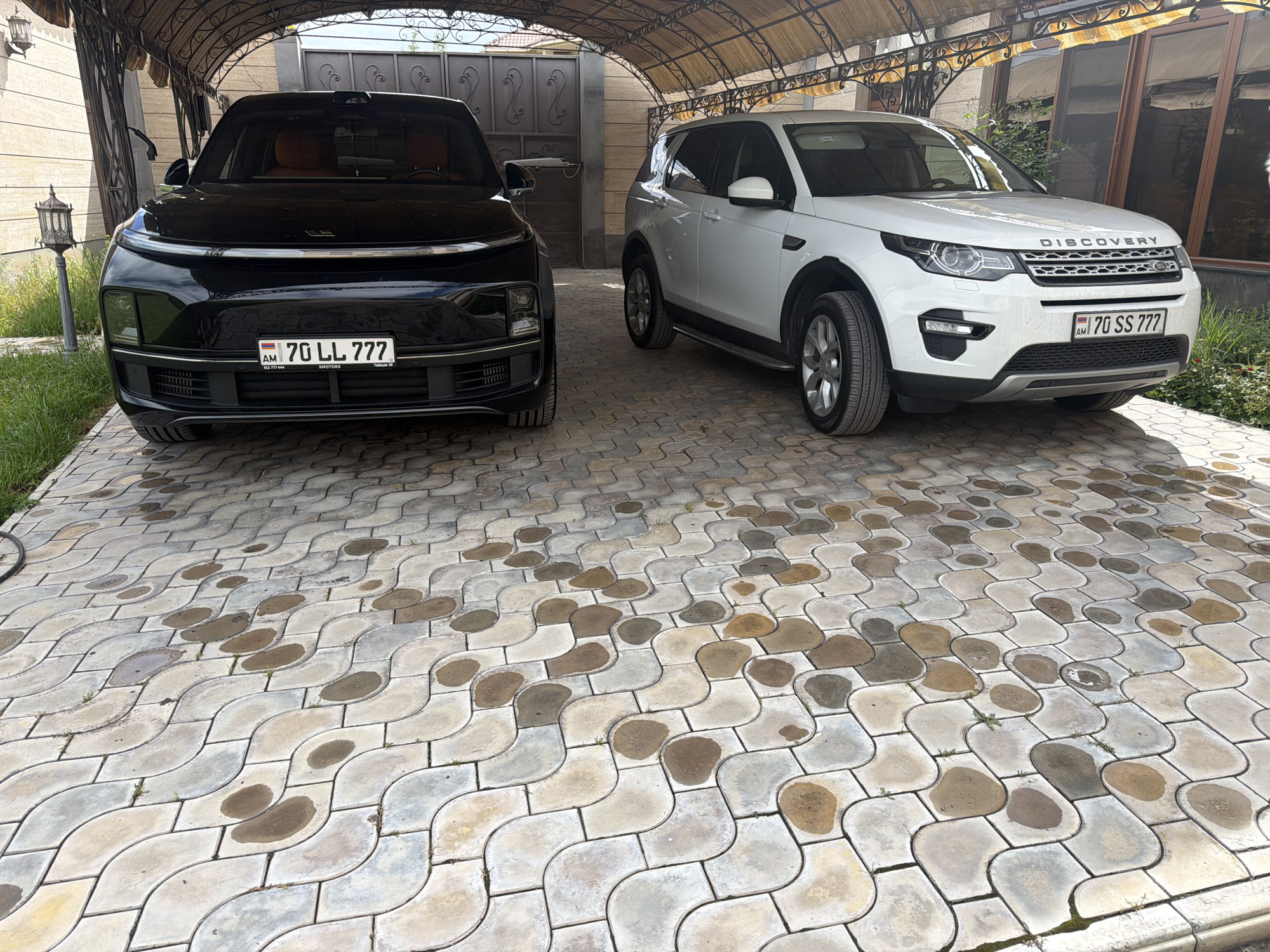 Two cars, a black vehicle with license plate "AM 70 LL 777" and a white "Land Rover Discovery" with license plate "AM 70 SS 777", are parked under a carport on a patterned stone driveway in front of a building. A lamppost stands in the yard on the left. The OCR text identifies "XMOTORS," and the license plates are Armenian. The location is likely in Armenia, potentially in or near the city of Dimitrov, Armenia.