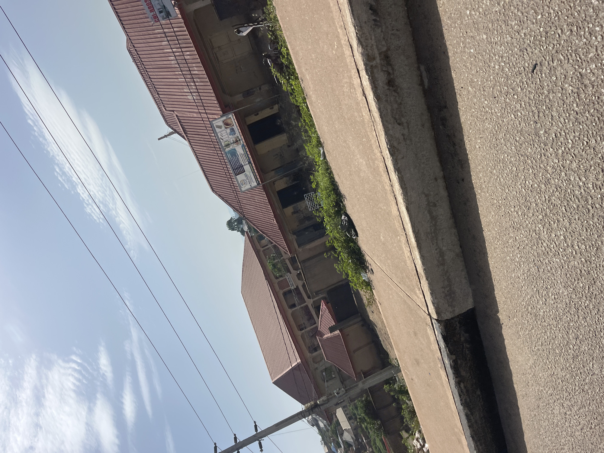 The image displays a street scene in Gura Top, Nigeria. The foreground is dominated by a paved road and a concrete curb, with shadows cast across the asphalt. In the mid-ground, a row of buildings with red metal roofs are visible. One building has a sign with text that appears to be in English, displaying images of what look like electronic components or devices. A utility pole with several wires runs along the left side of the frame. The background shows more buildings and vegetation on a slight incline under a cloudy sky. No people or overt actions are depicted.