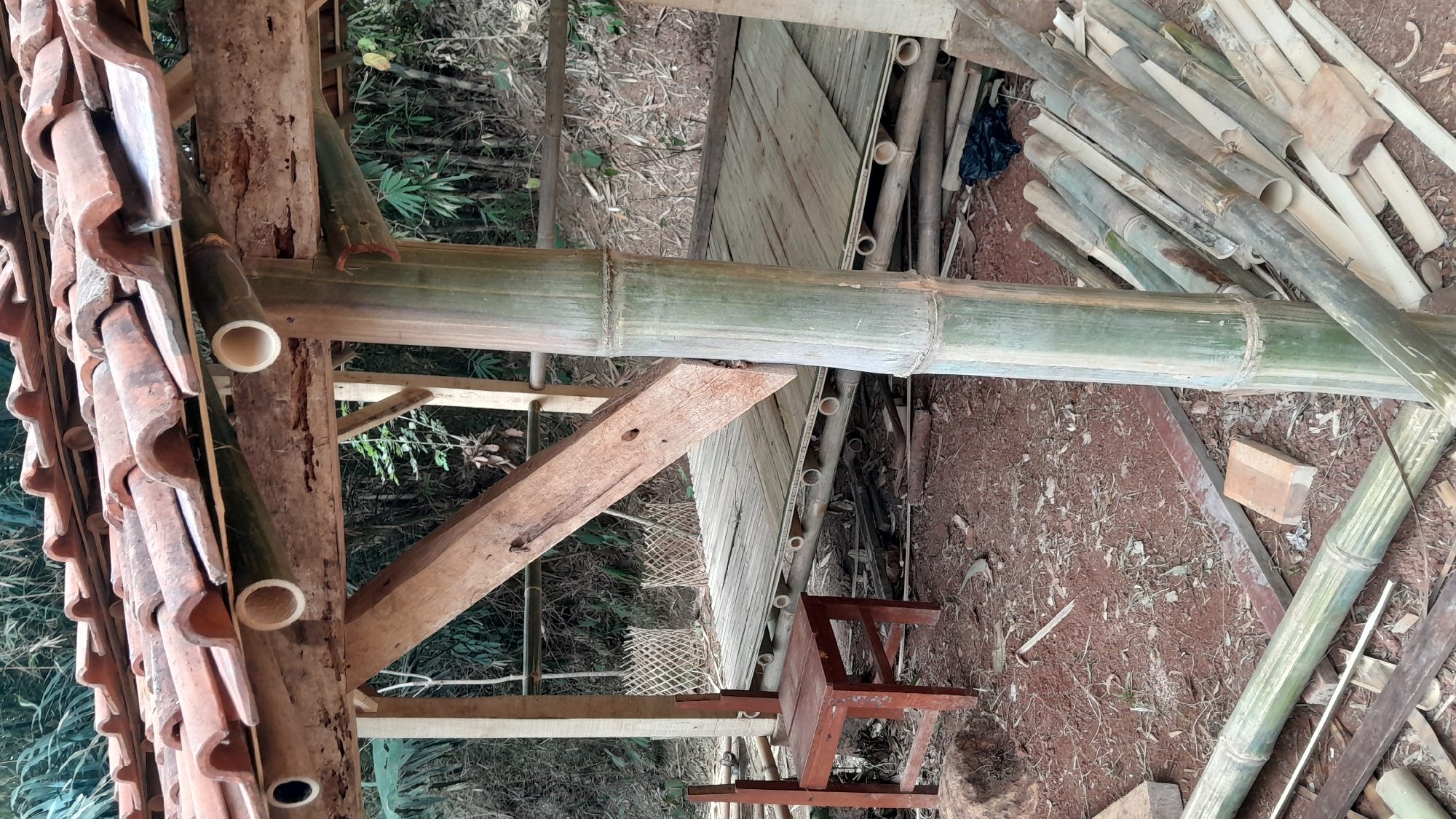 The image depicts an unfinished outdoor structure, likely a gazebo or small pavilion, constructed primarily from bamboo and wood. The roof is made of terracotta tiles, supported by wooden beams and bamboo poles. A wooden chair sits on the dirt ground to the left of the structure, near a partially constructed wooden platform. The ground is covered in red soil and scattered debris, including more bamboo poles, wood planks, and a black plastic bag. In the background, a dense forest of green foliage is visible. The scene suggests a rural or semi-rural setting, possibly in a village or natural area. Given the context of Sumedang, Indonesia, this location would likely be an outdoor area within the regency, surrounded by its characteristic tropical vegetation.