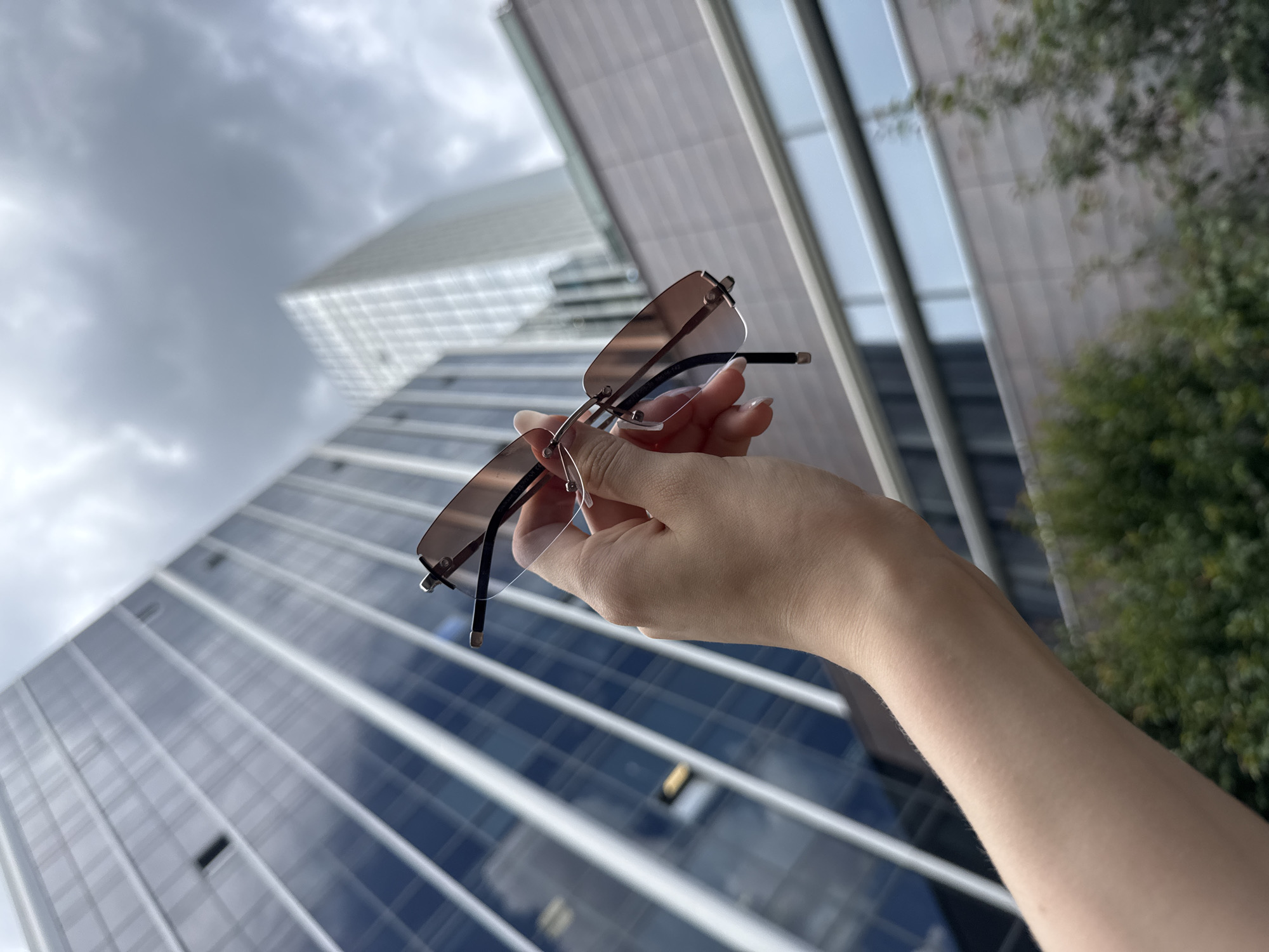 An over-the-shoulder shot shows a hand holding rimless sunglasses with rose-tinted lenses. The hand, with manicured nails, is positioned in the lower right quadrant of the frame. The background is dominated by the reflective glass facade of a modern skyscraper, its vertical lines creating a sense of depth and scale. The sky above is overcast with gray clouds. The image appears to be taken in an urban environment, likely in the city of Yekaterinburg, Russia, given the architectural style of the building.