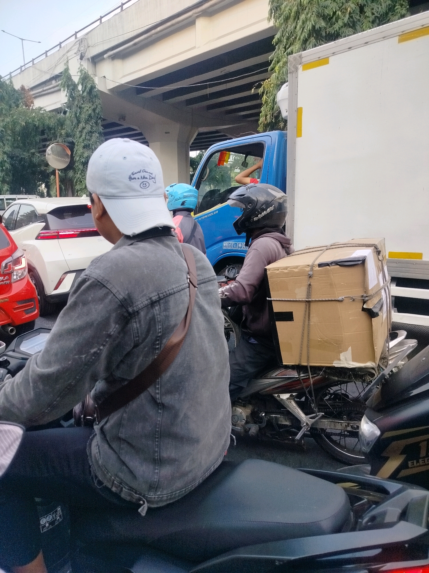 In East Jakarta, Indonesia, a traffic jam is depicted under an elevated road. In the foreground, a person wearing a denim jacket and a white baseball cap is seated on a motorcycle, facing away from the camera. Behind them, another motorcyclist wearing a black helmet and a gray hoodie is also on a motorcycle, carrying a large cardboard box secured with ropes. To the right, a blue truck is visible, and to the left, a white car and a red car are part of the traffic congestion. The environment suggests a typical urban traffic scene with overhead structures and vegetation.