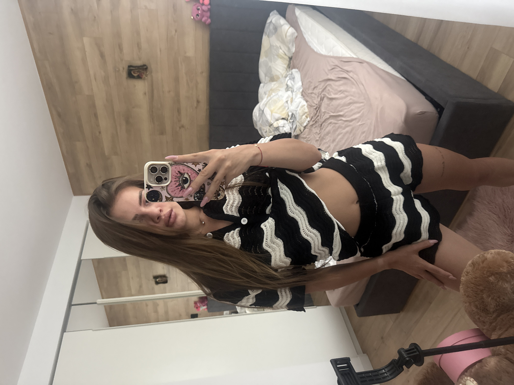 The image displays a mirror selfie of a young woman taken in a bedroom, oriented sideways. The woman is positioned horizontally across the frame, holding a smartphone with a distinctive pink case featuring a large eye design. She has long brown hair and is wearing a black and white striped knitted or crocheted top and matching shorts. Her fingernails are painted a light pink shade, and she has a small red string bracelet on her left wrist and a delicate necklace. Her expression is neutral.
The background, reflected in the mirror, shows a room with light wooden flooring visible on the left and right due to the image's rotation. A bed with a dark grey headboard and frame occupies the right side of the frame, covered with a pink/beige fitted sheet and a white patterned duvet. A small pink plush toy is visible on the floor near the top left corner. In the bottom right, a large brown plush teddy bear and a black tripod or stand are partially visible. A white wardrobe or closet with reflective doors is seen along the far left edge of the image. There is no clearly legible text.