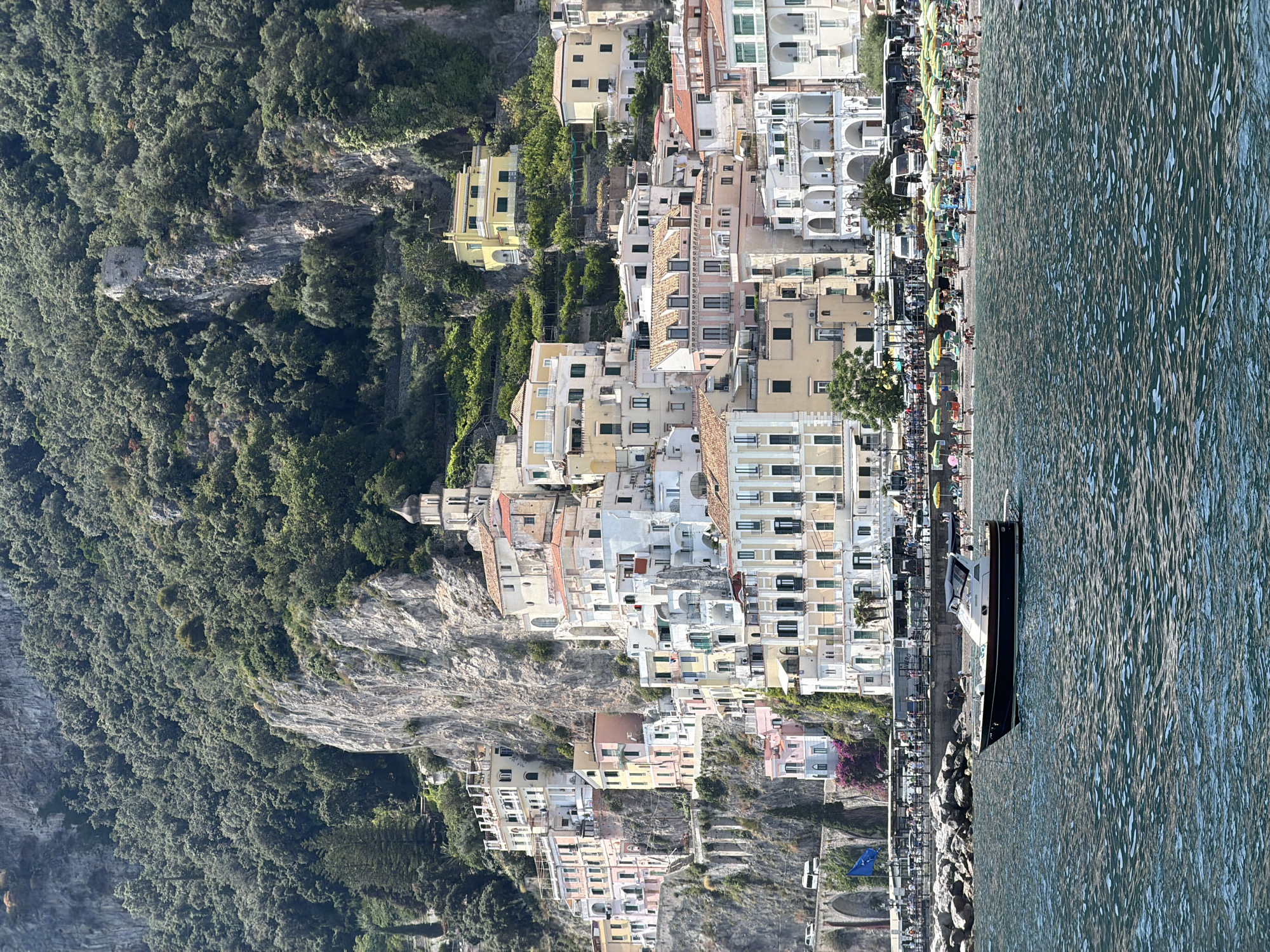 The image shows a coastal town built on a steep hillside. The town is densely populated with buildings of varying heights and colors, mostly pastel shades, constructed on the rocky terrain. Lush green vegetation covers the hillside to the left, contrasting with the built environment.
To the right, the water of the sea is visible, with a boat anchored near the shore. Along the waterfront, there is a narrow beach with numerous people and umbrellas.
The location is likely Pogerola, Italy, due to the characteristic architecture and coastal setting. The image captures the integration of a built environment with a natural landscape, a typical feature of the Amalfi Coast.