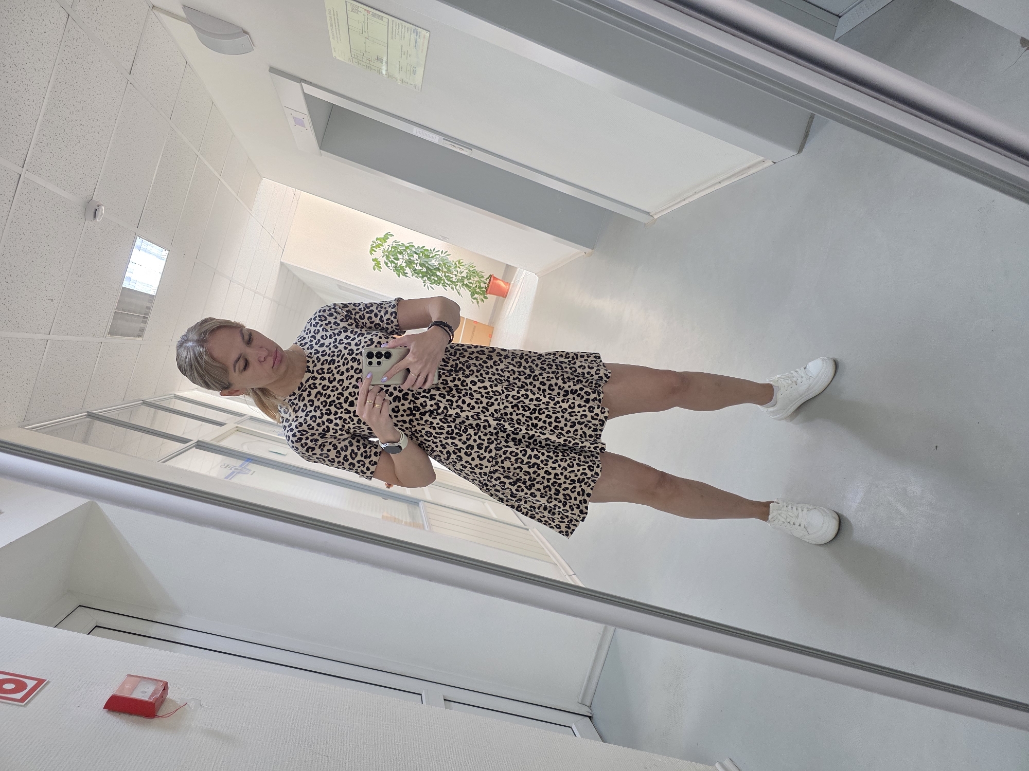 In a well-lit, indoor hallway, a woman takes a mirror selfie. She is wearing a short-sleeved, leopard-print dress that ends above her knees, paired with white lace-up sneakers. Her blonde hair is pulled back, and she is holding a smartphone in her right hand, pointing it towards the mirror. A large mirror dominates the left side of the frame, reflecting the woman and the hallway. The hallway features a grey floor, white walls, and a textured white ceiling with recessed lighting. To the right of the woman, a potted green plant is visible in front of a light-colored wall. Further down the hallway, an elevator door is apparent. The overall scene suggests an office building or a similar professional environment, consistent with locations in Chaikovsky, Russia.