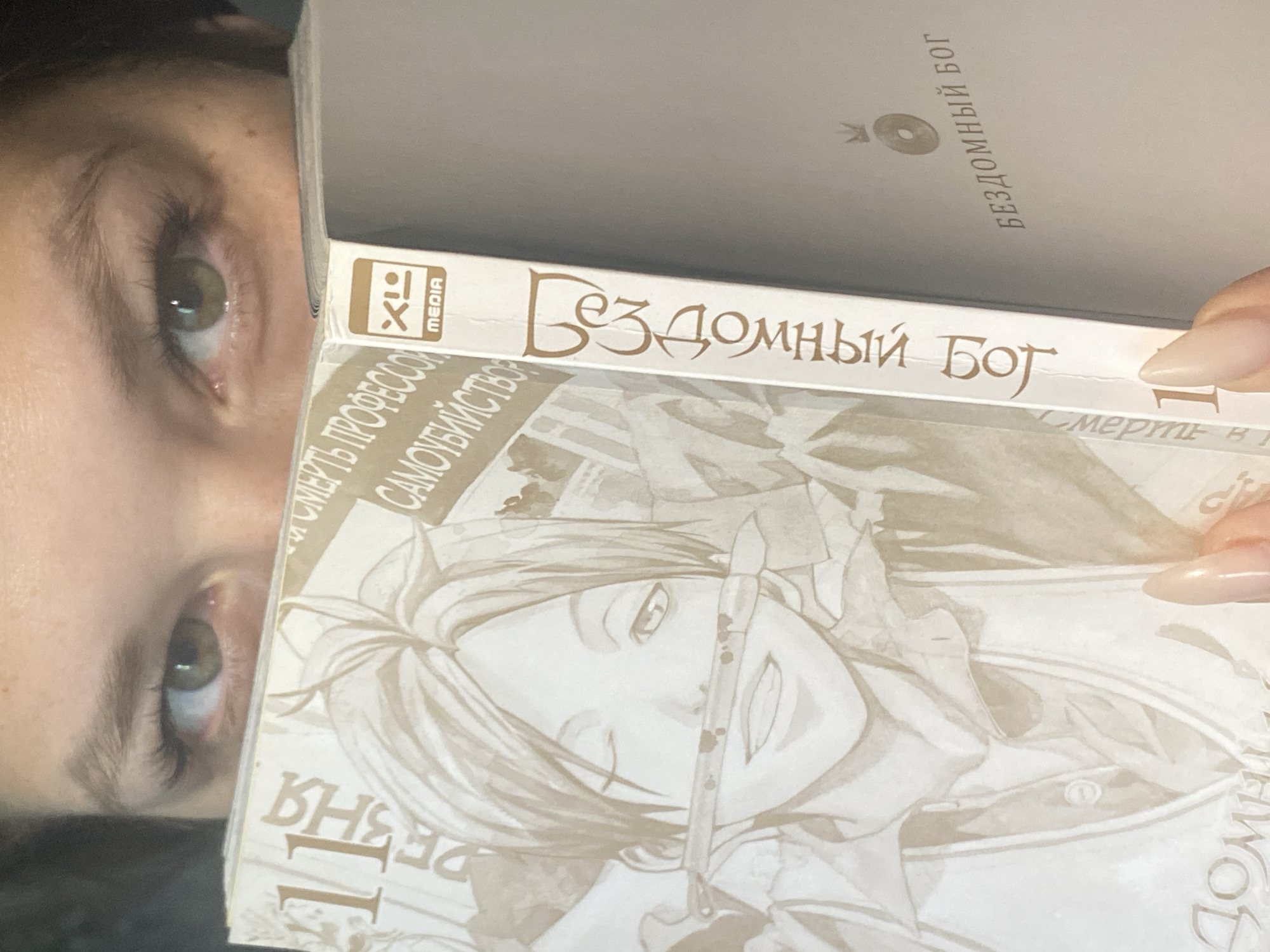 A close-up, off-kilter photograph shows a person holding two books in front of their face, partially obscuring it. The books appear to be manga or graphic novels, written in Russian, and have a stylized cover with a drawing of a character. The top book is a light color with the title "Бездомный Бог" ("Homeless God"). The bottom book also has the same title "Бездомный Бог", as well as "СМЕРТЬ ПРОФЕССОР СУИЦИДОВ" which roughly translates to "Death of Suicide Professor". The background is out of focus and unclear, but it is most likely taken indoors in an unknown location; there's no information that can be used to determine that it was taken in Chaikovsky, Russia. The person's eyes are visible above the books. They have light skin and light eyes. Their nails are long and polished. The lighting in the photo appears to be somewhat dim.