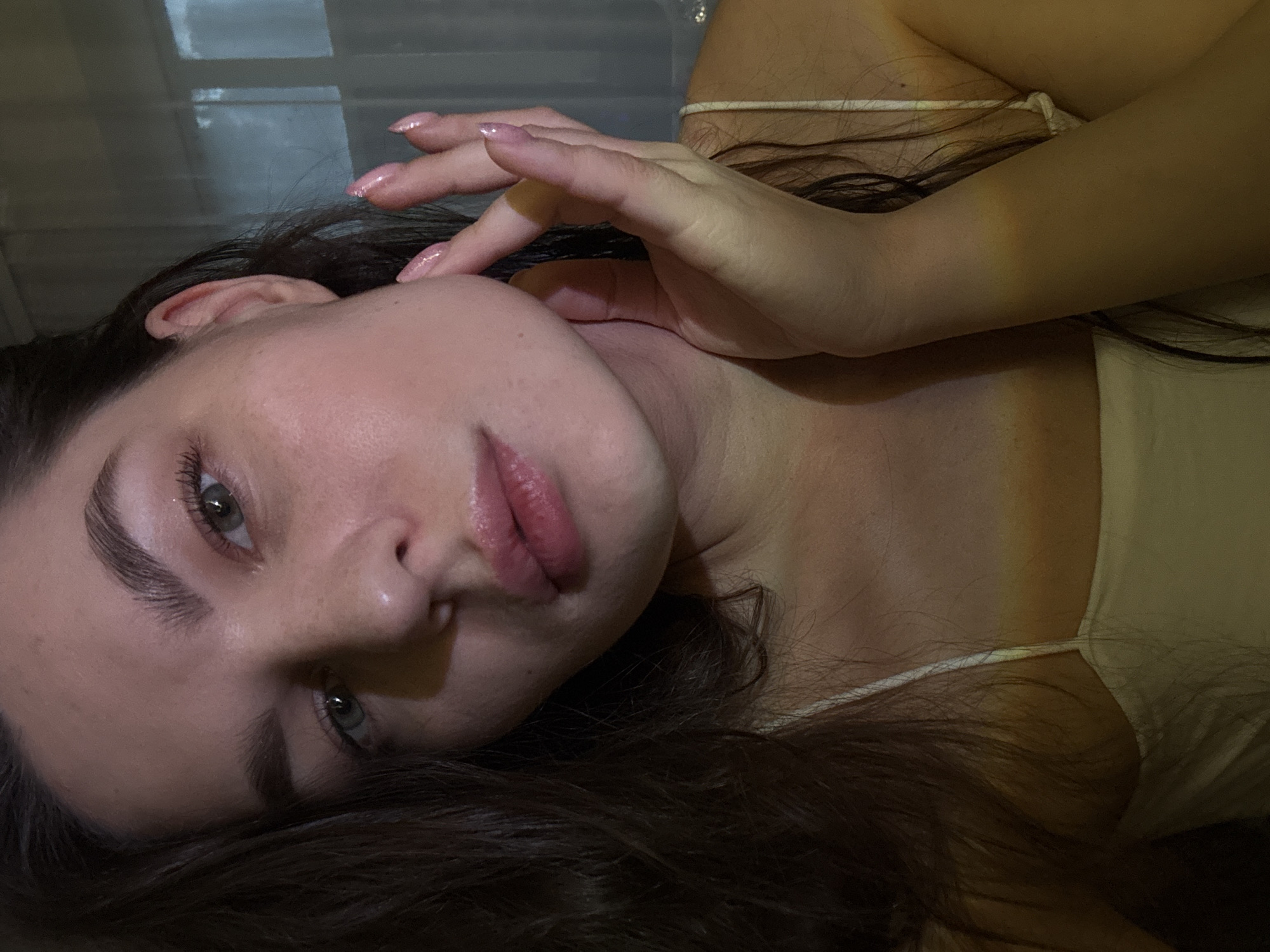 A close-up, shoulder-up, portrait-style photograph shows a young woman with dark hair and light skin. She is looking directly at the camera with a soft expression. Her right hand is raised, with her fingers gently touching her cheek and jawline. Her nails are painted with a light, shimmery pink polish. She is wearing a cream-colored camisole. The background appears to be indoors with diffused lighting, possibly near a window with sheer curtains, suggesting a residential setting. No specific landmarks or details are visible to indicate a location within Chaikovsky, Russia.