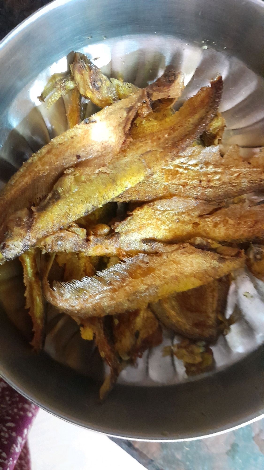 A close-up, overhead shot shows a metallic bowl filled with golden-brown, fried fish. The fish appear to be small, whole fish that have been seasoned and fried until crispy. They are arranged in a layered fashion within the bowl, with some pieces overlapping others. The bowl itself has a slightly worn, reflective surface, showing subtle ripples from its manufacturing. The background is blurred, but suggests a table surface and a hint of a patterned cloth on the left. The lighting is bright, casting a warm glow on the food. The scene appears to be in a domestic setting within the city of Bhubaneswar Municipal Corporation, India, possibly a kitchen or dining area, where a meal is being prepared or served.