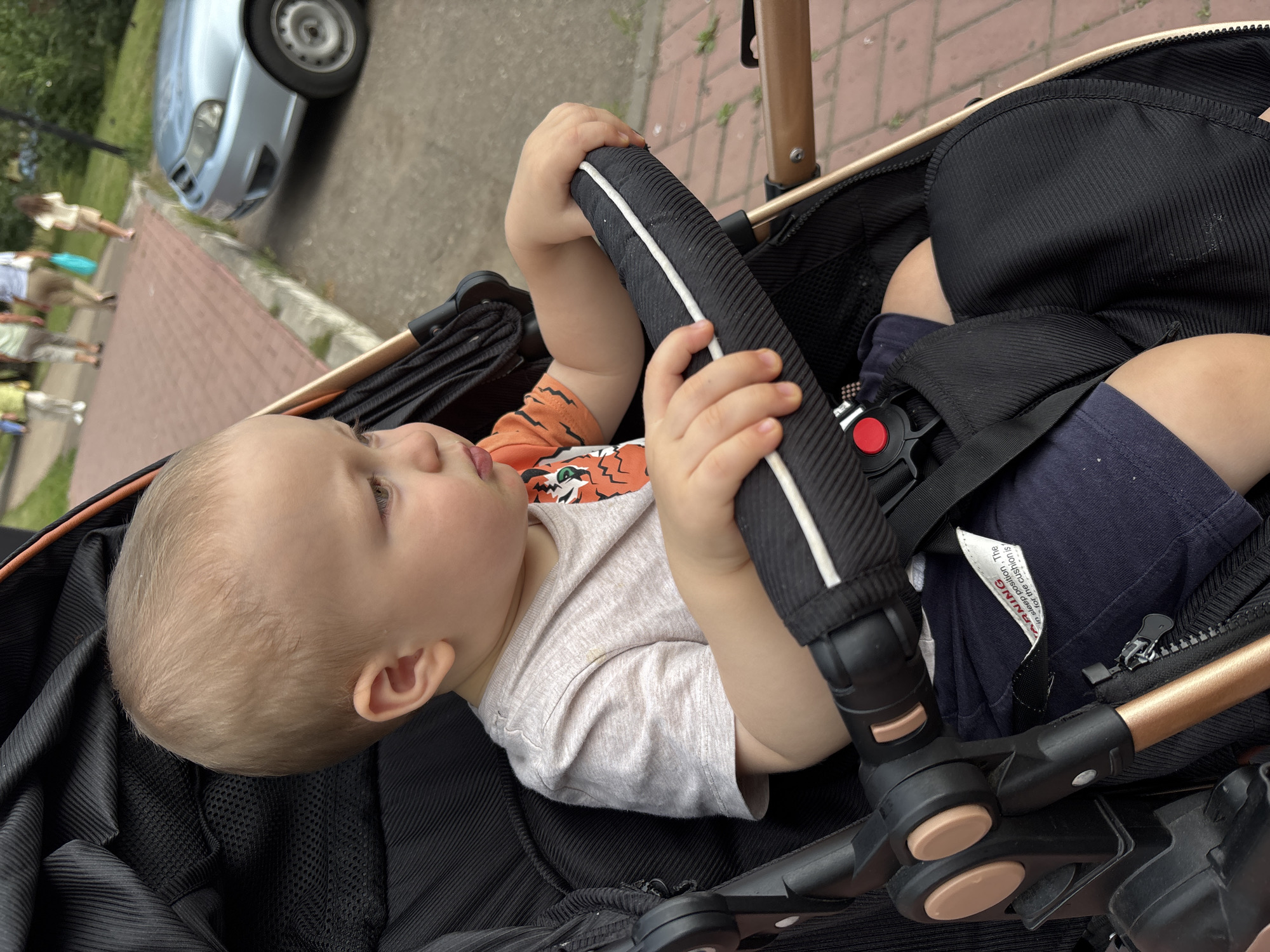 A toddler sits in a black stroller, grasping the handlebar. The child wears a light-colored t-shirt with an orange tiger graphic on the sleeve and dark blue shorts. In the background, people walk along a paved path, and a silver car is parked nearby. The scene appears to be in an outdoor urban area, possibly a park or sidewalk in Naberezhnye Chelny, Russia.