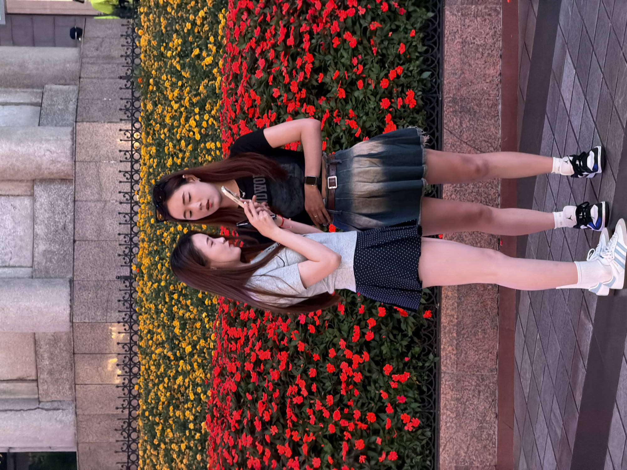 Two young women stand in front of a vibrant floral display in Yuzhong District, China. The woman on the left, wearing a grey t-shirt and a black polka dot skirt, is looking down at her phone. The woman on the right, in a black t-shirt and a faded denim skirt, is also looking at her phone, with one hand on the other woman's arm. The floral display features rows of red and yellow flowers. Behind the flowers, a large stone structure with columns is visible, suggesting a public monument or building. The ground is paved with square tiles.