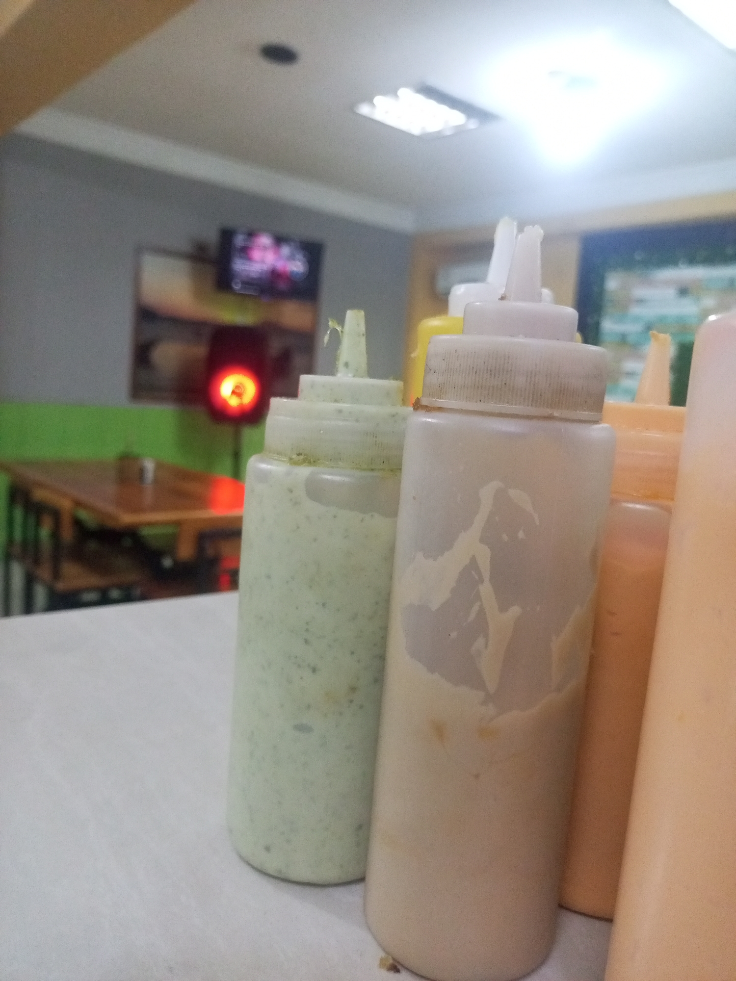 The image shows several squeeze bottles filled with sauces or condiments on a white table. The bottles are various colors, including light green, off-white, yellow, and peach. The bottles are slightly dirty with dried sauce residue.
In the blurred background, there is a wooden table and chairs. Further in the background, there is a wall painted green and gray and a television mounted on the wall. The location appears to be a restaurant or eating establishment in Juan Griego, Venezuela.