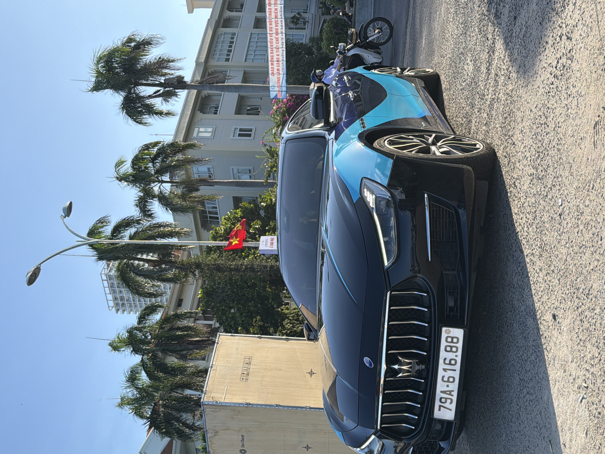 The image captures a black Maserati Ghibli with a custom blue and black wrap parked on a street in Nha Trang, Vietnam. The car's license plate reads "79A-616.88." In the background, palm trees and modern buildings are visible, suggesting an urban coastal environment. A Vietnamese flag is displayed on a pole. A banner with Vietnamese text, likely advertising an event or message, is hung across the street. A person on a motorcycle, wearing a helmet, is seen to the right of the car. The overall scene is illuminated by bright sunlight, indicating daytime.