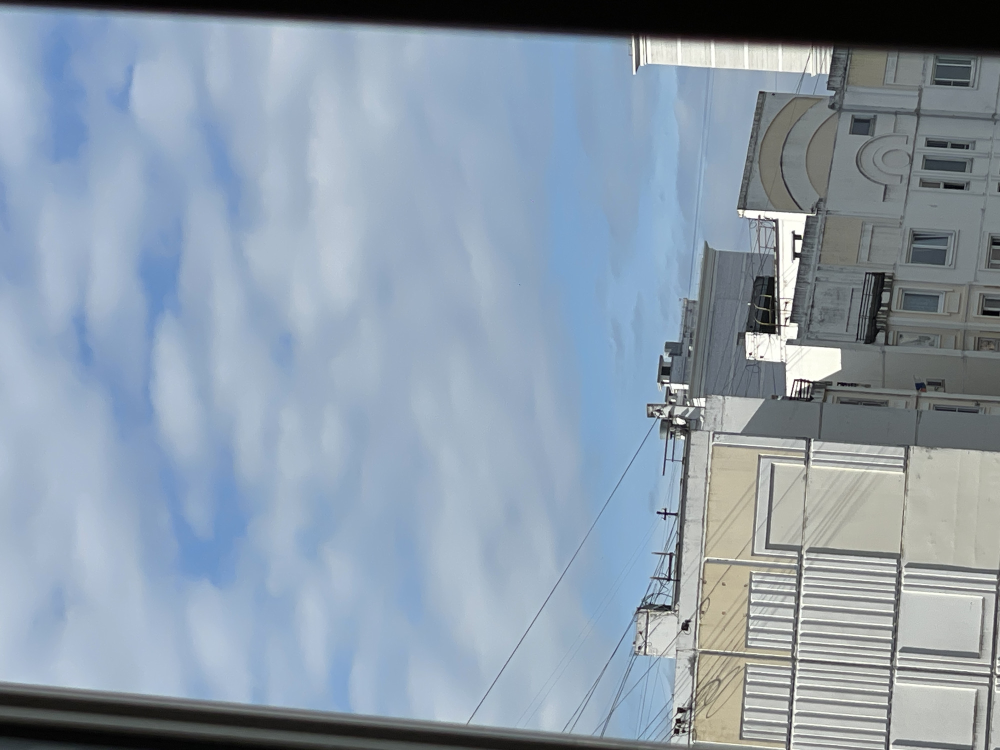 The image shows a view of several multi-story buildings against a partly cloudy sky. The buildings have varied architectural styles, with some featuring ribbed facade details and decorative elements. Power lines are visible against the sky. The overall scene suggests an urban environment. There are no people or animals visible in the image. The lighting indicates it is daytime. The provided crops focus on details of the sky and the buildings, highlighting the textures and architectural features.