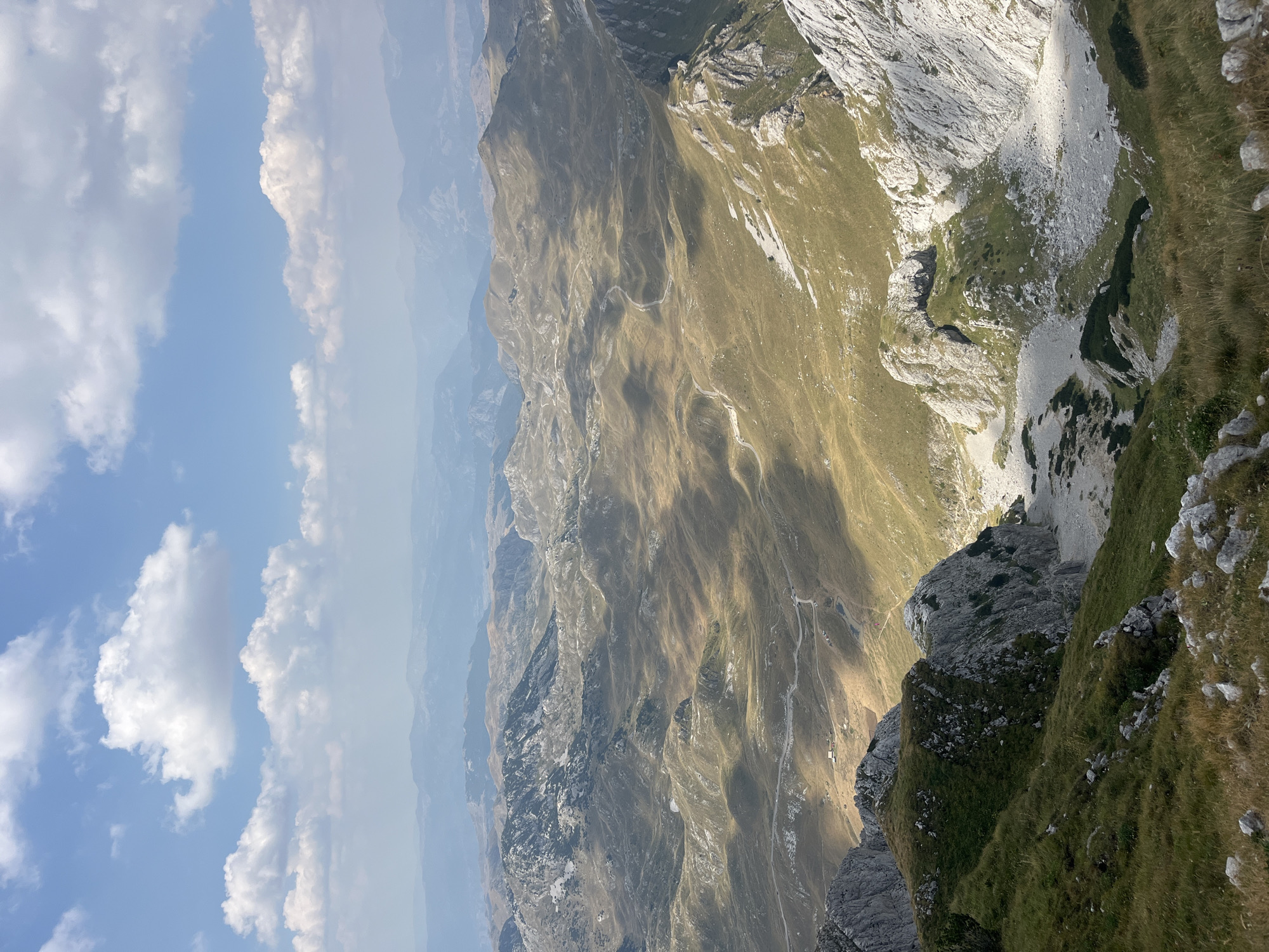 The image is a high-angle, wide shot of a rugged mountain landscape with rolling hills and rocky outcrops. The terrain is covered in dry, golden grass, interspersed with patches of green vegetation and bare rock. A winding road is visible snaking through the valleys, with a few small buildings or structures dotted along its path. In the foreground, the sharp, rocky edge of a cliff descends into a valley. The background shows a series of progressively distant mountain ranges fading into a hazy blue. The sky is a bright blue with scattered white cumulus clouds, some of which cast shadows on the mountains below. The lighting suggests it is daytime, with sunlight illuminating the scene from the left, creating strong contrasts between light and shadow. The scene depicts a remote, natural environment, characteristic of a mountainous region. The location is identified as Боричје, Montenegro, which is consistent with the rugged and expansive mountainous terrain depicted.